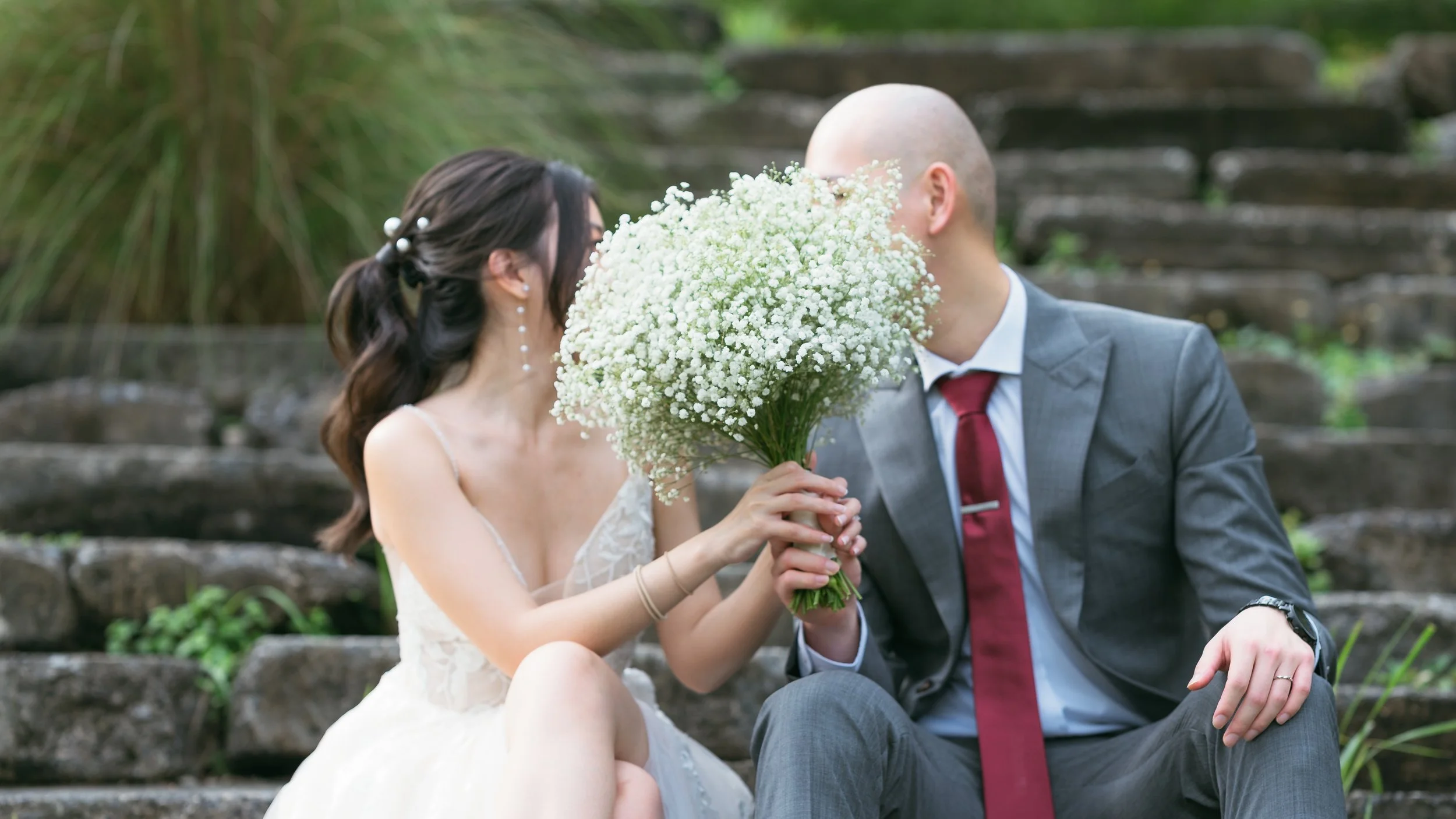 A bride and groom sitting on stone steps outdoors, kissing behind a large bouquet of white baby's breath flowers, with greenery in the background.