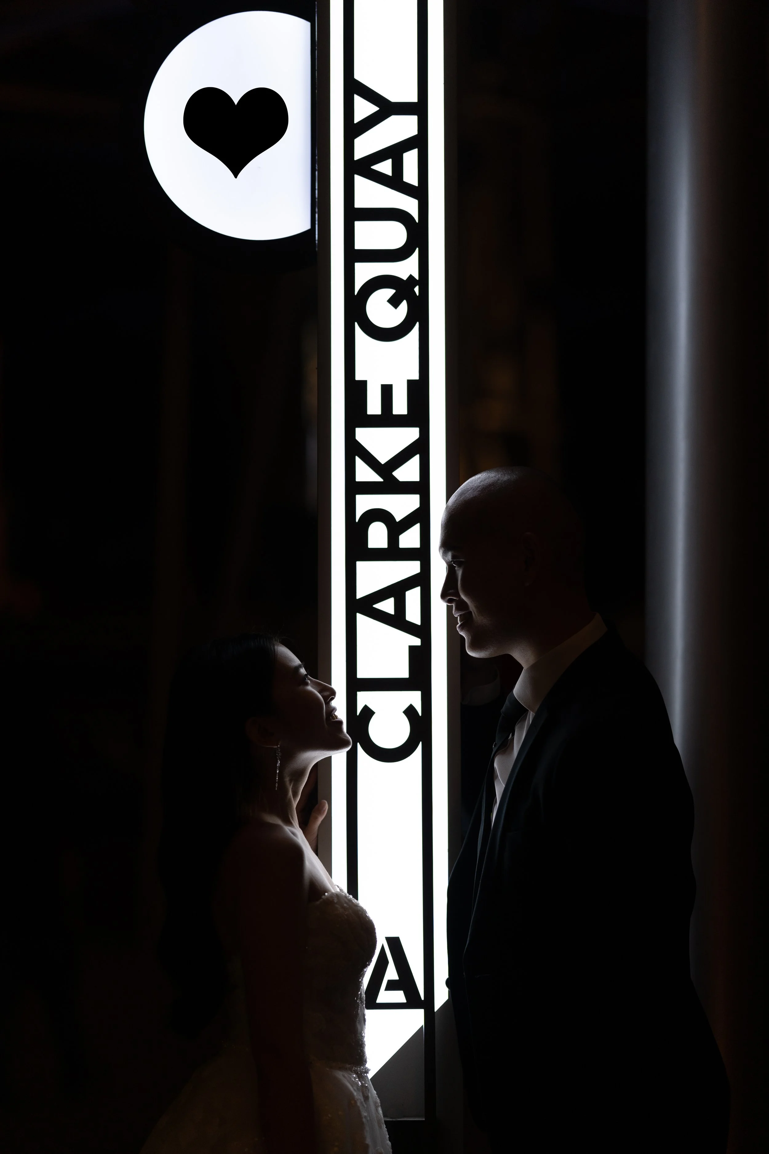 Silhouettes of a bride and groom facing each other in front of a backlit sign with a heart logo, on their wedding day.