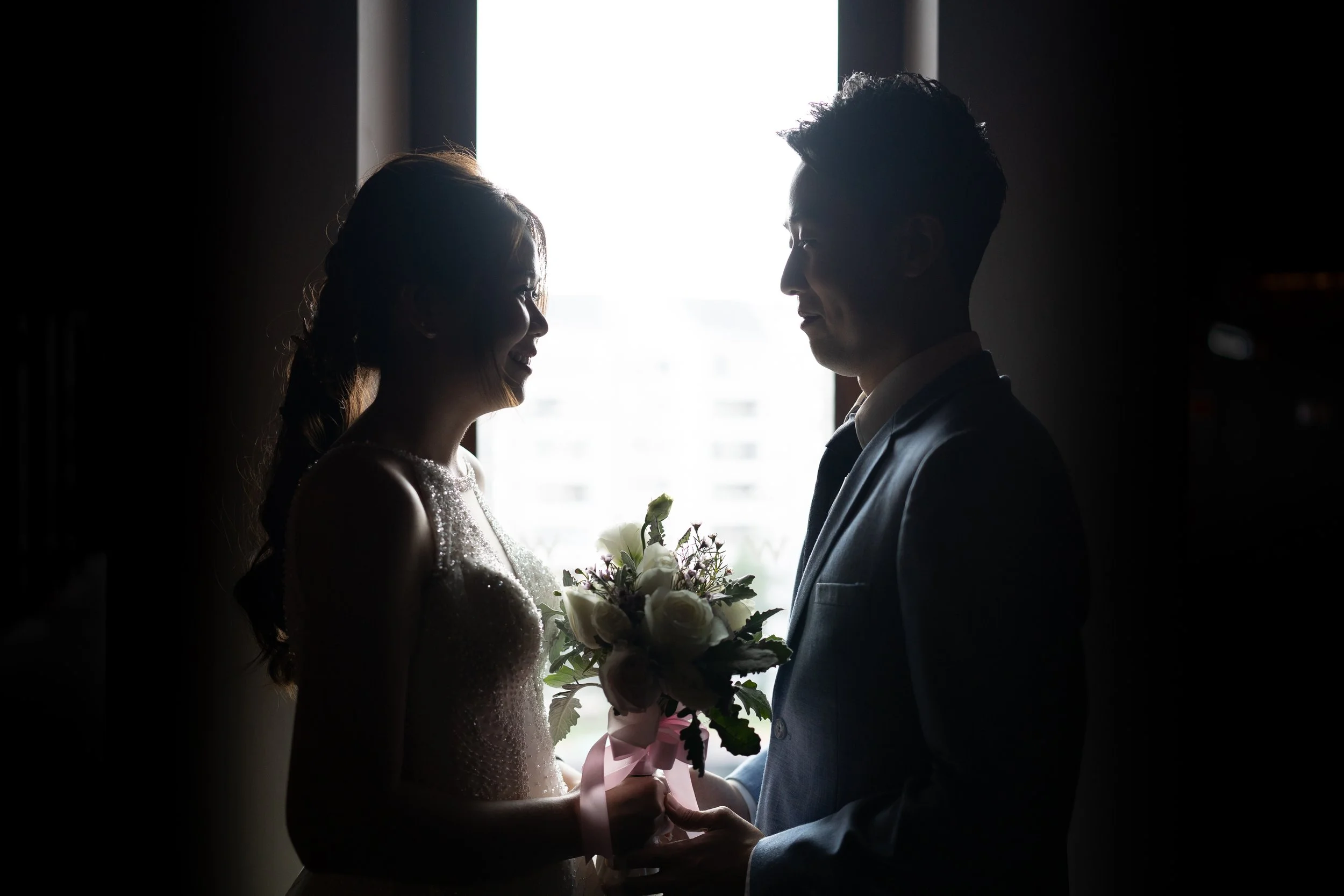 Silhouette of a bride and groom holding hands near a window, with the bride holding a bouquet of white flowers and pink ribbon.