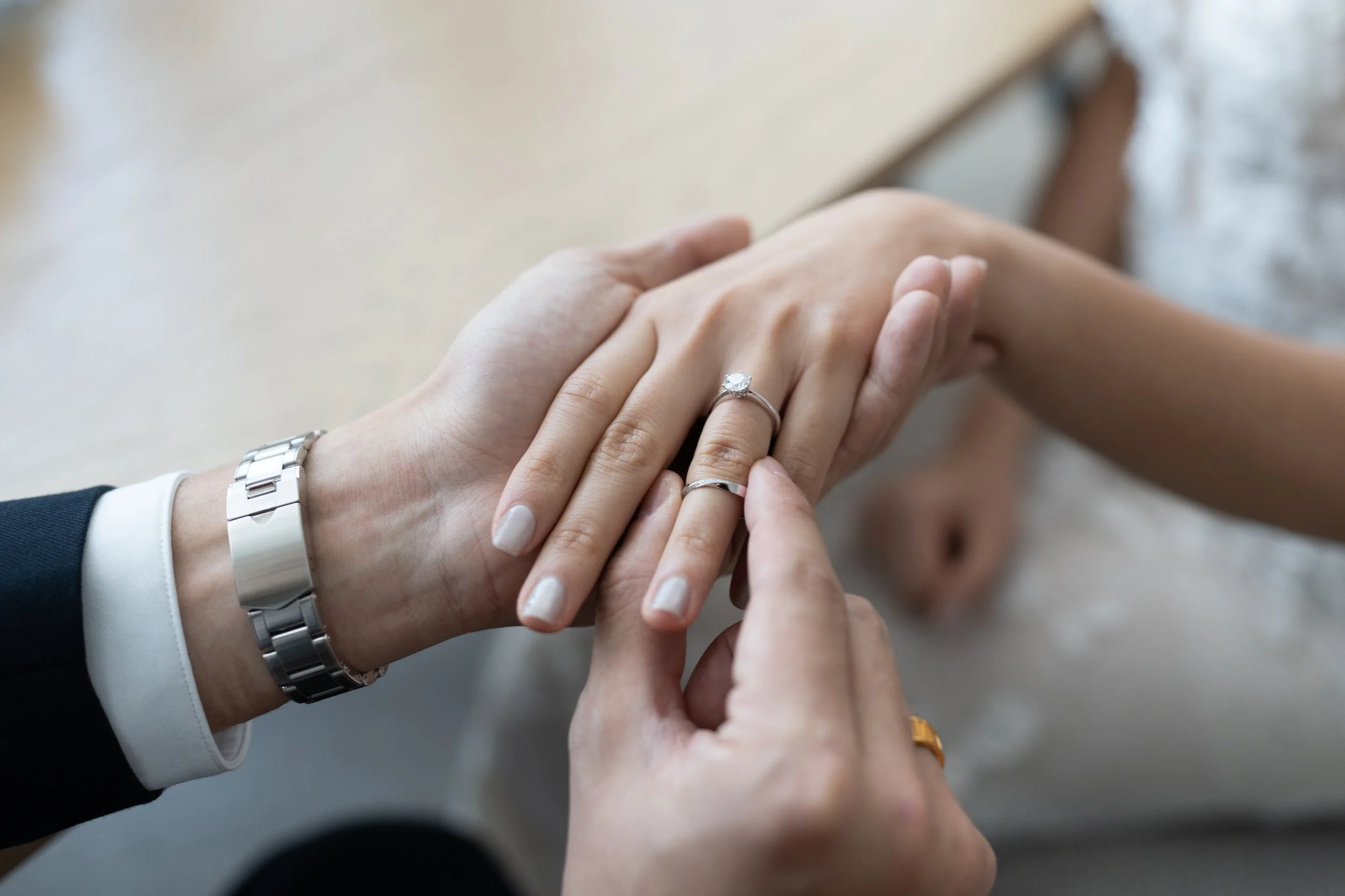 Close-up of a couple's hands. The man is placing an engagement ring on the woman's finger, and they are both wearing wedding bands. The woman has a diamond ring, and the man has a metal watch on his wrist.