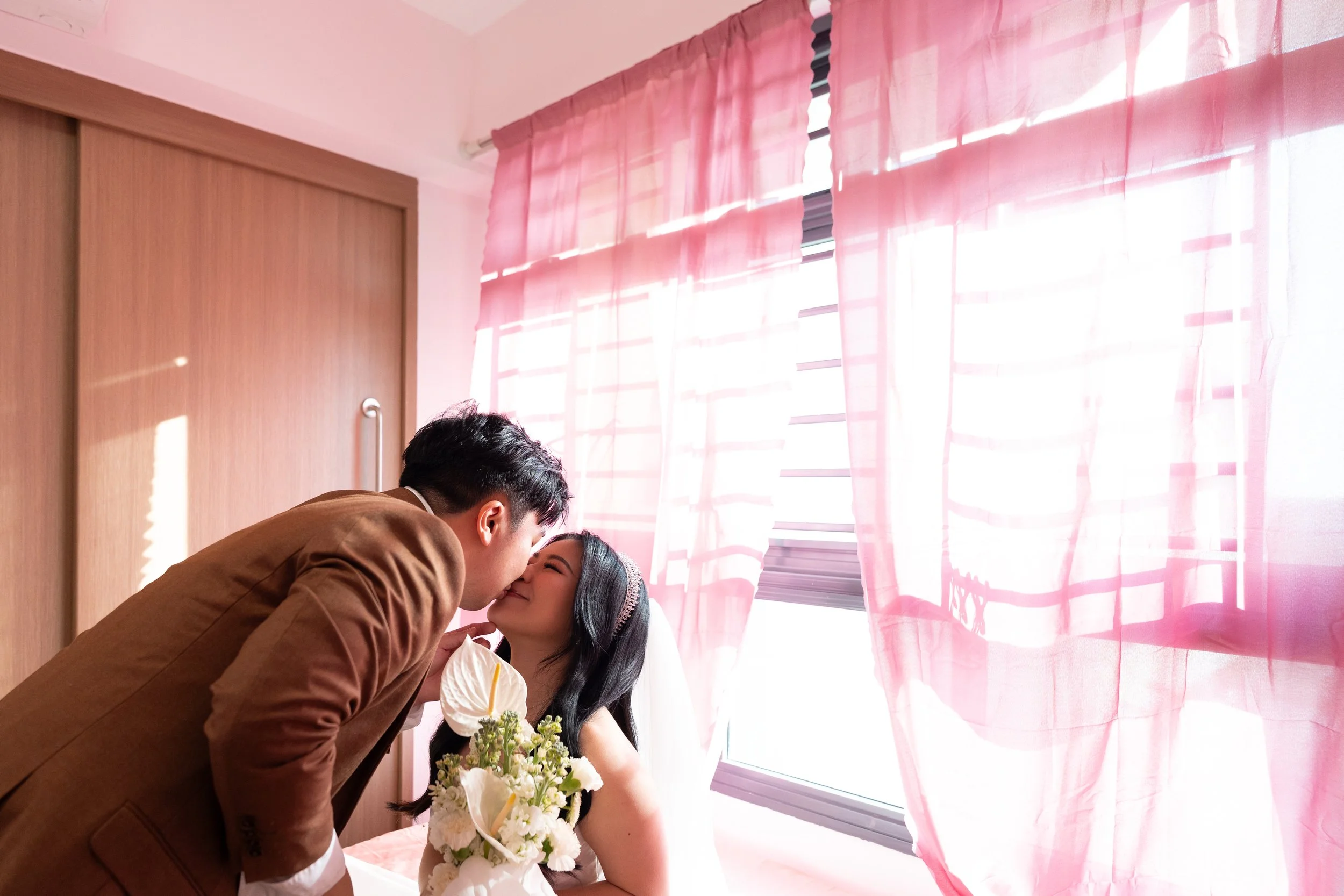 A couple sharing a kiss at a wedding, with the bride holding a bouquet of white flowers, in a room with pink curtains and sunlight filtering through the window.