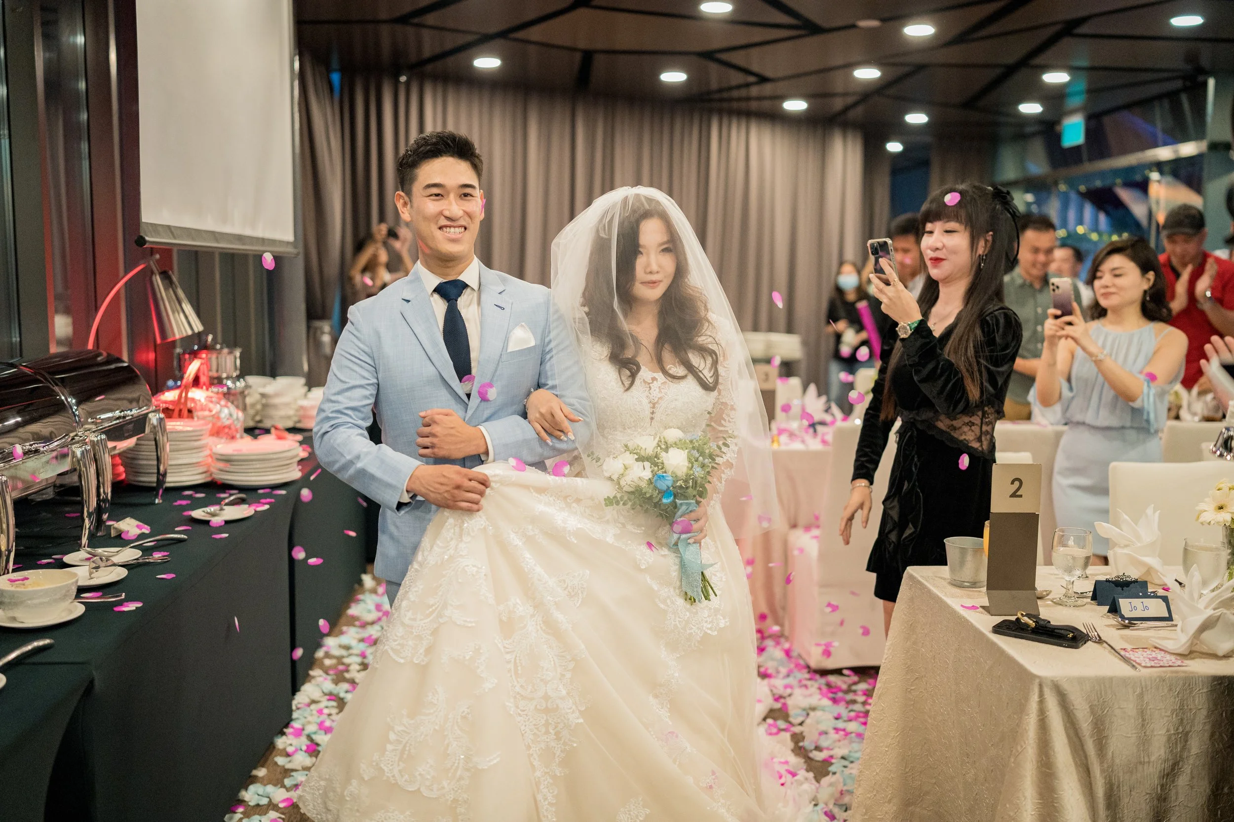 A bride and groom walk arm-in-arm at their wedding reception, surrounded by guests taking photos and throwing pink and white confetti, with a decorated banquet hall in the background.