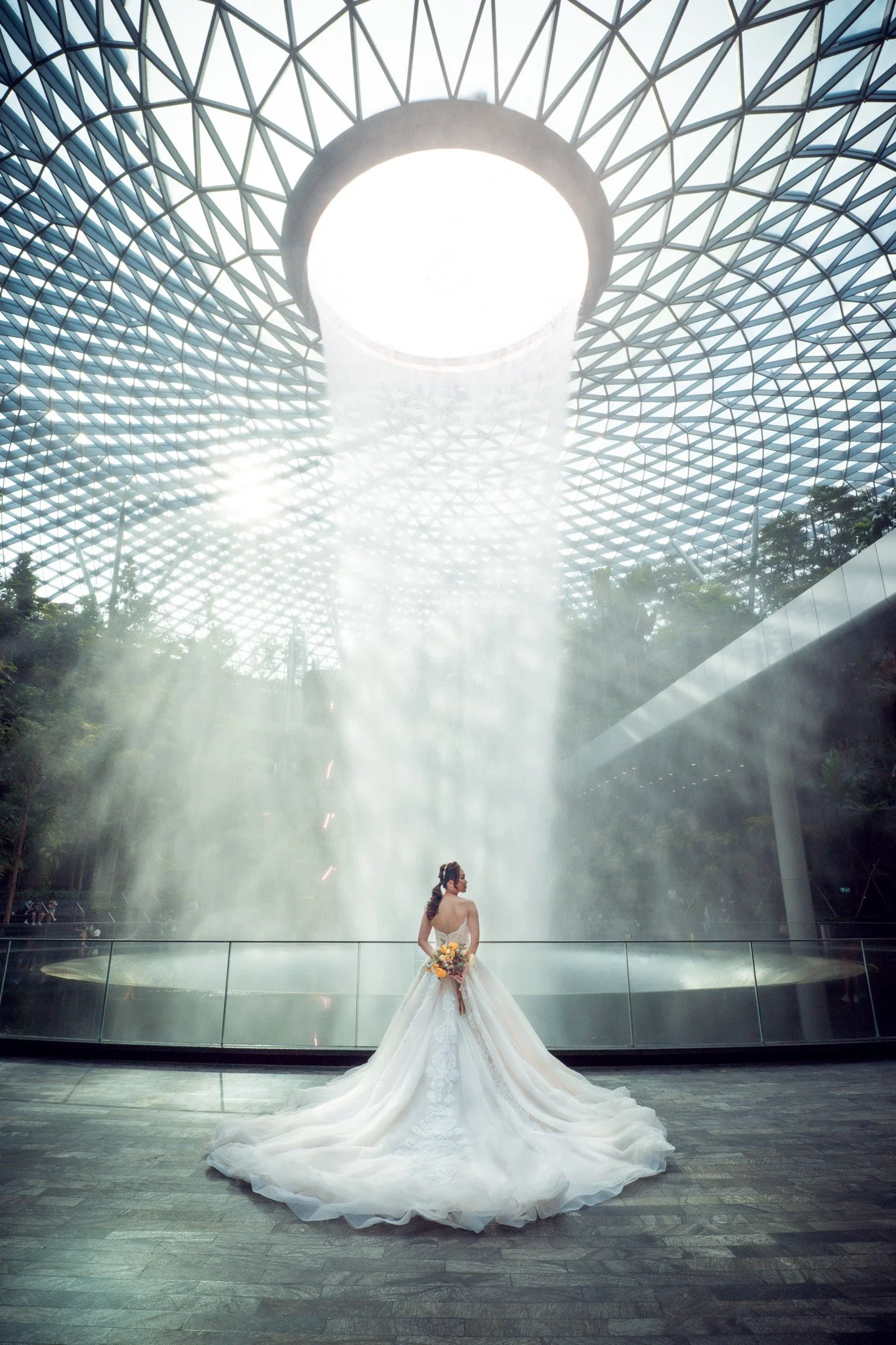 A woman in a wedding dress holding a bouquet, standing in front of a large waterfall inside a modern glass and metal structure with a circular opening in the roof, allowing sunlight to stream down.