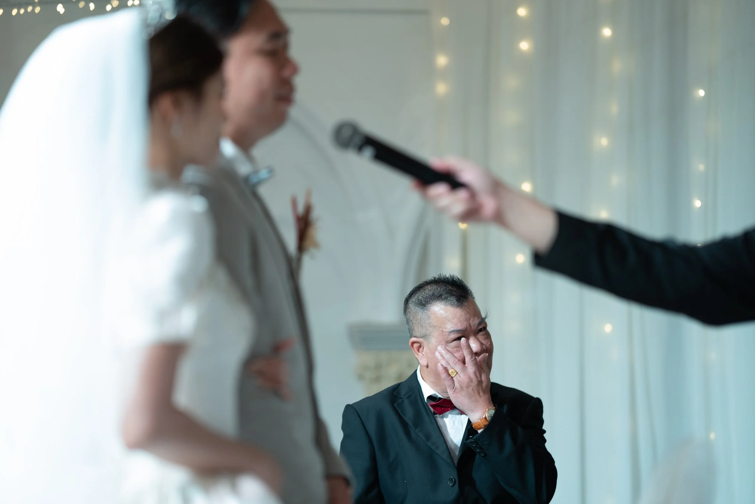 A man in a tuxedo with a red bow tie and a woman in a wedding dress standing in front of a man in a suit with a boutonniere, while someone off-camera is holding a microphone near the man.