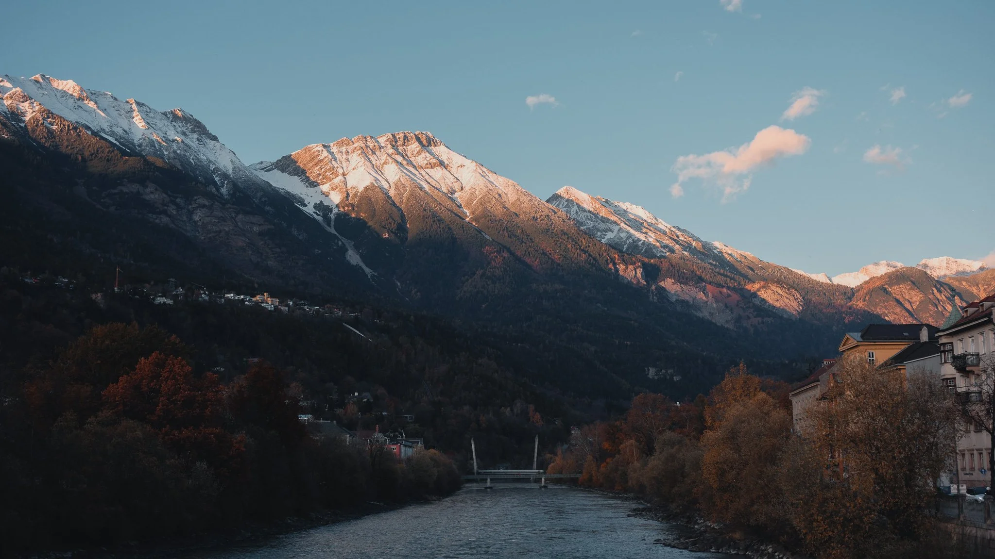 A scenic view of snow-capped mountains with a river running through a town in the foreground, autumn trees along the riverbank, and a bridge crossing the river.