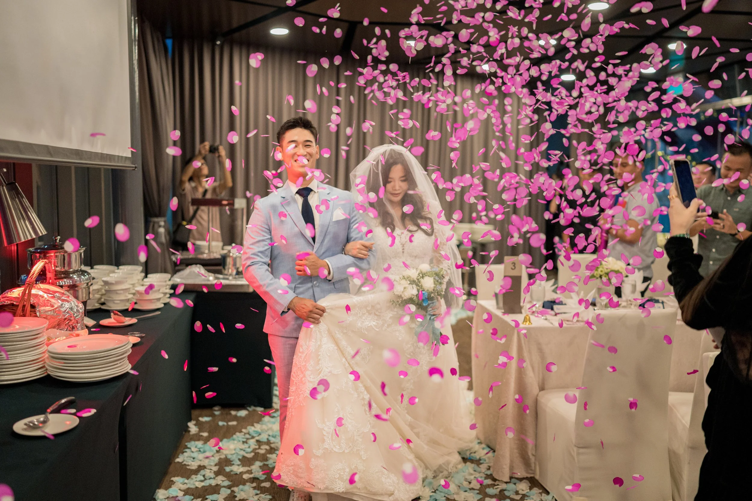 A bride and groom celebrating their wedding reception with pink confetti falling around them. The groom is smiling while the bride is holding a bouquet. Guests are taking photos in the background.