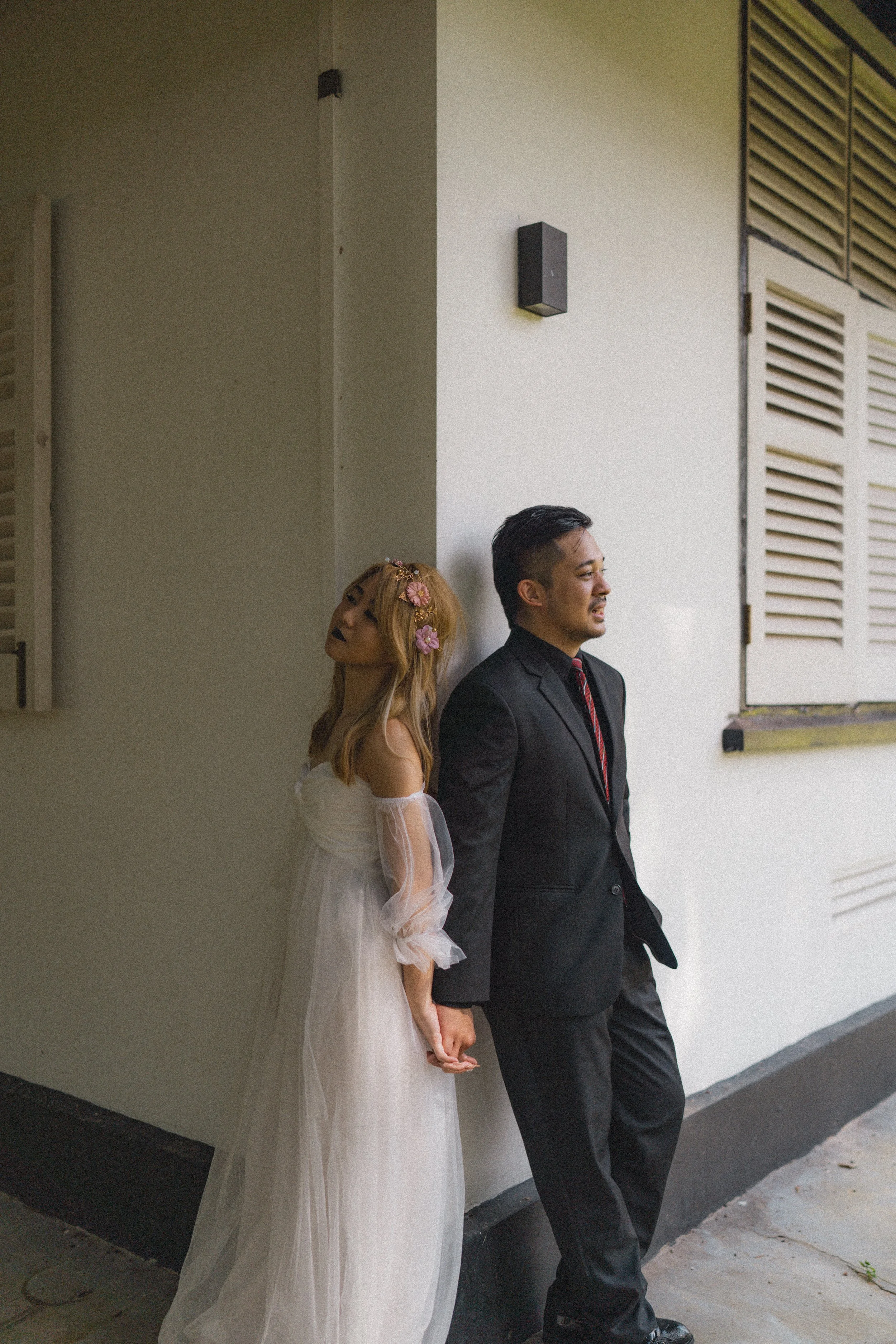 A couple dressed formally, the woman in a white dress with floral hair accessories and the man in a dark suit, stand back-to-back leaning against a wall, holding hands, outside near a building with white walls and shutters.
