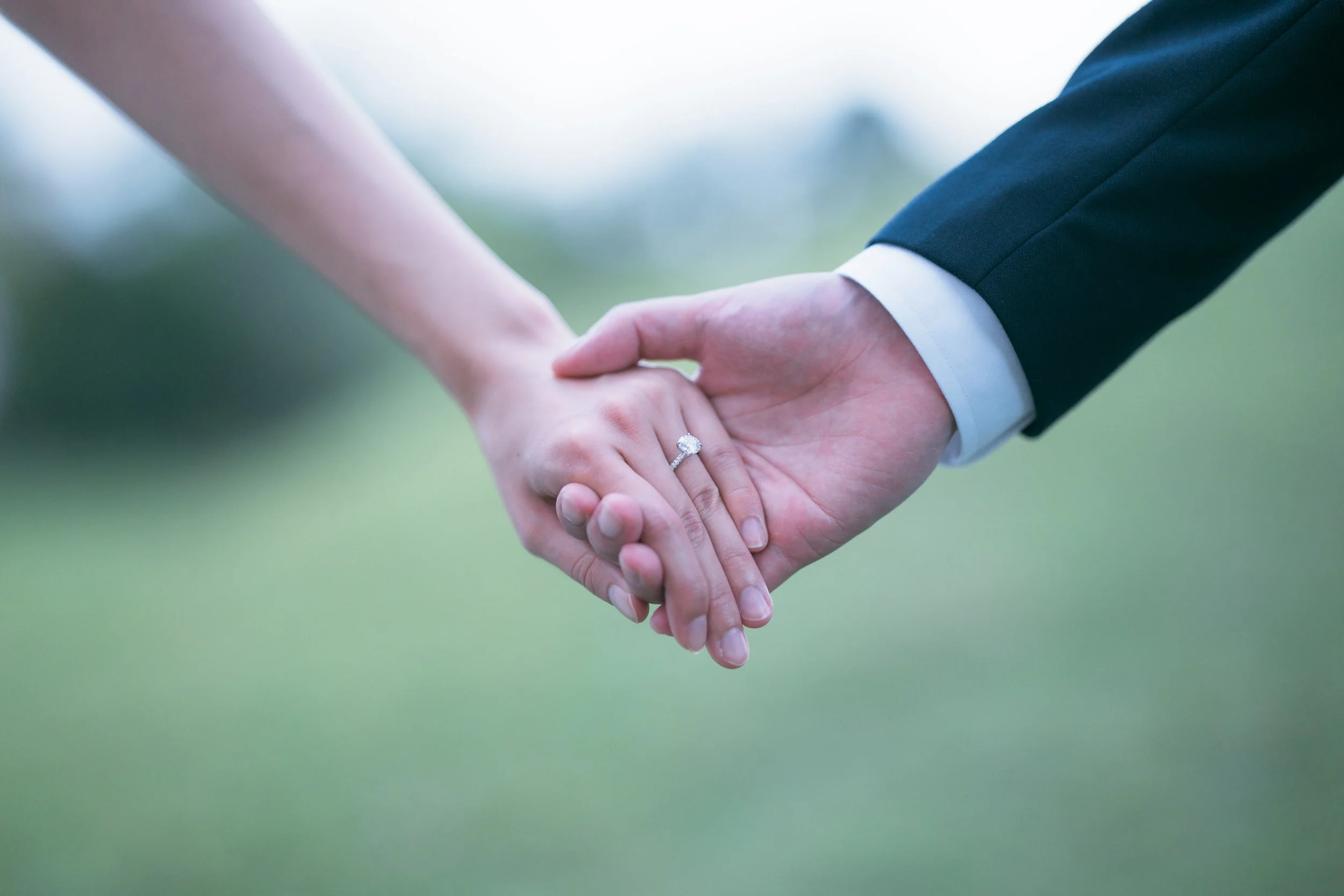 Close-up of a man and woman holding hands outdoors; the woman is wearing an engagement ring.