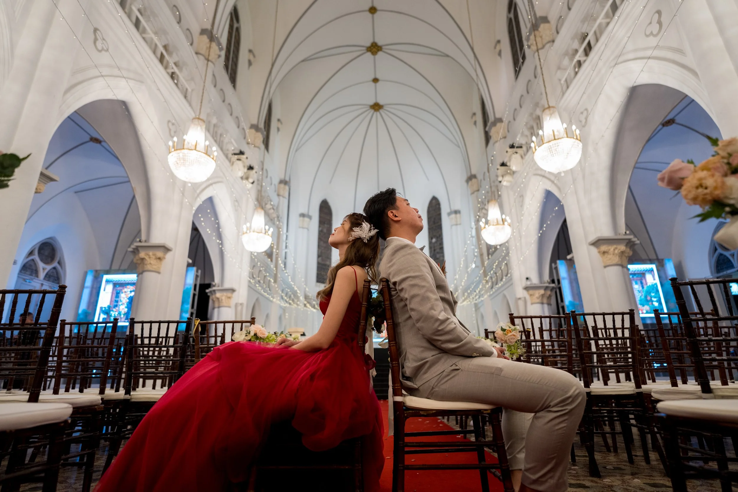 A couple sitting back to back on chairs inside a church, in a wedding setting with elegant chandeliers and string lights, with flowers on their chairs.