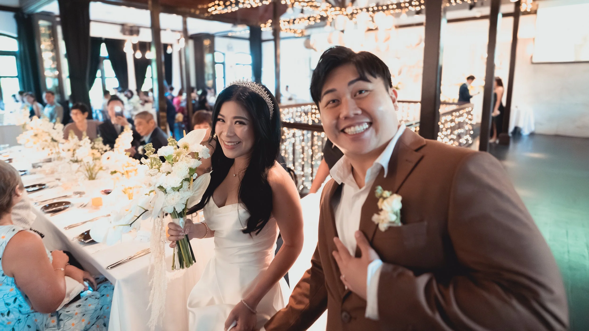 A bride and groom smiling at a wedding reception, with guests seated at a decorated table in the background.