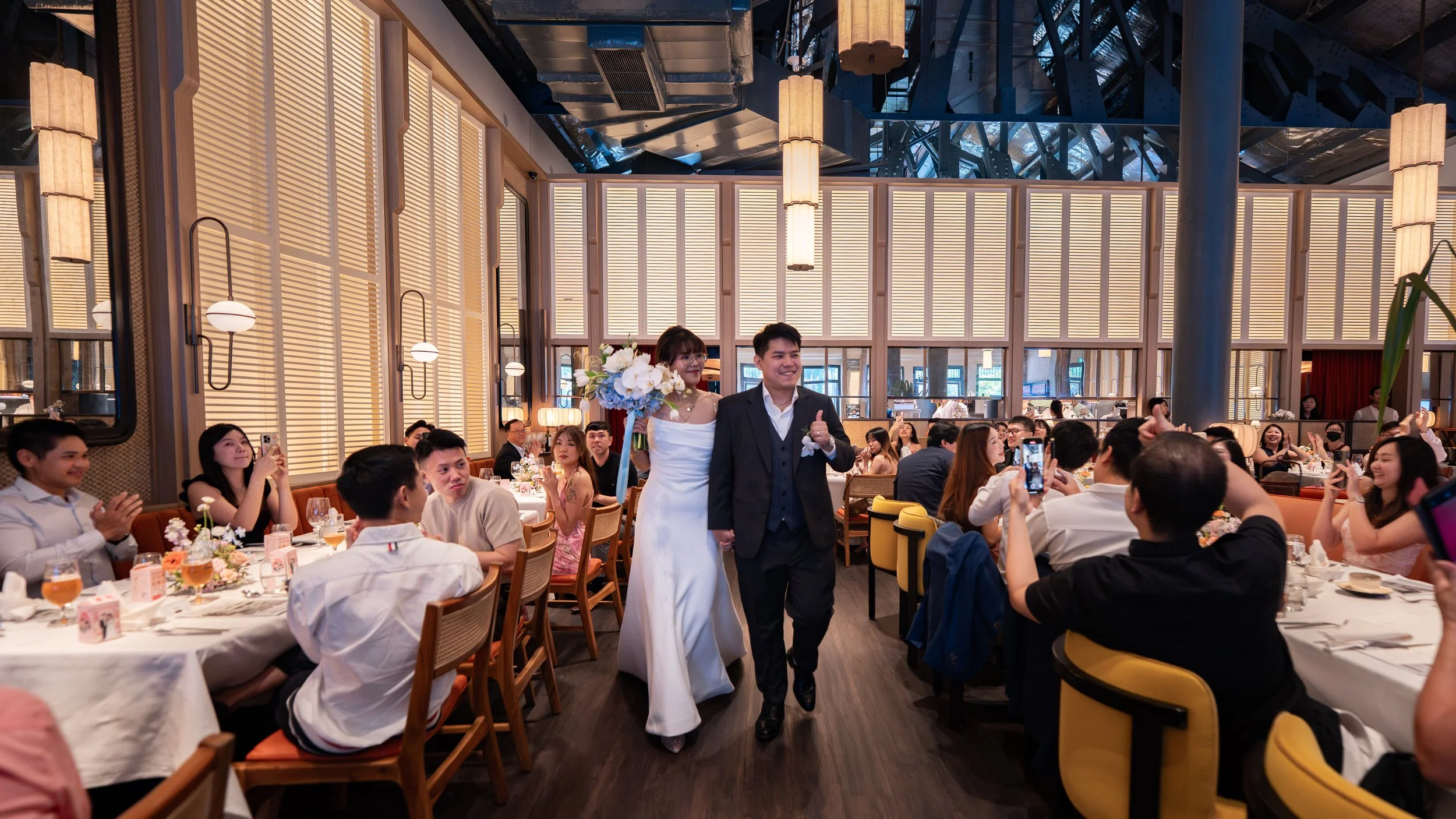 A wedding reception with a bride in a white dress and groom in a suit walking down the aisle, surrounded by seated guests taking photos and clapping, in a decorated banquet hall.