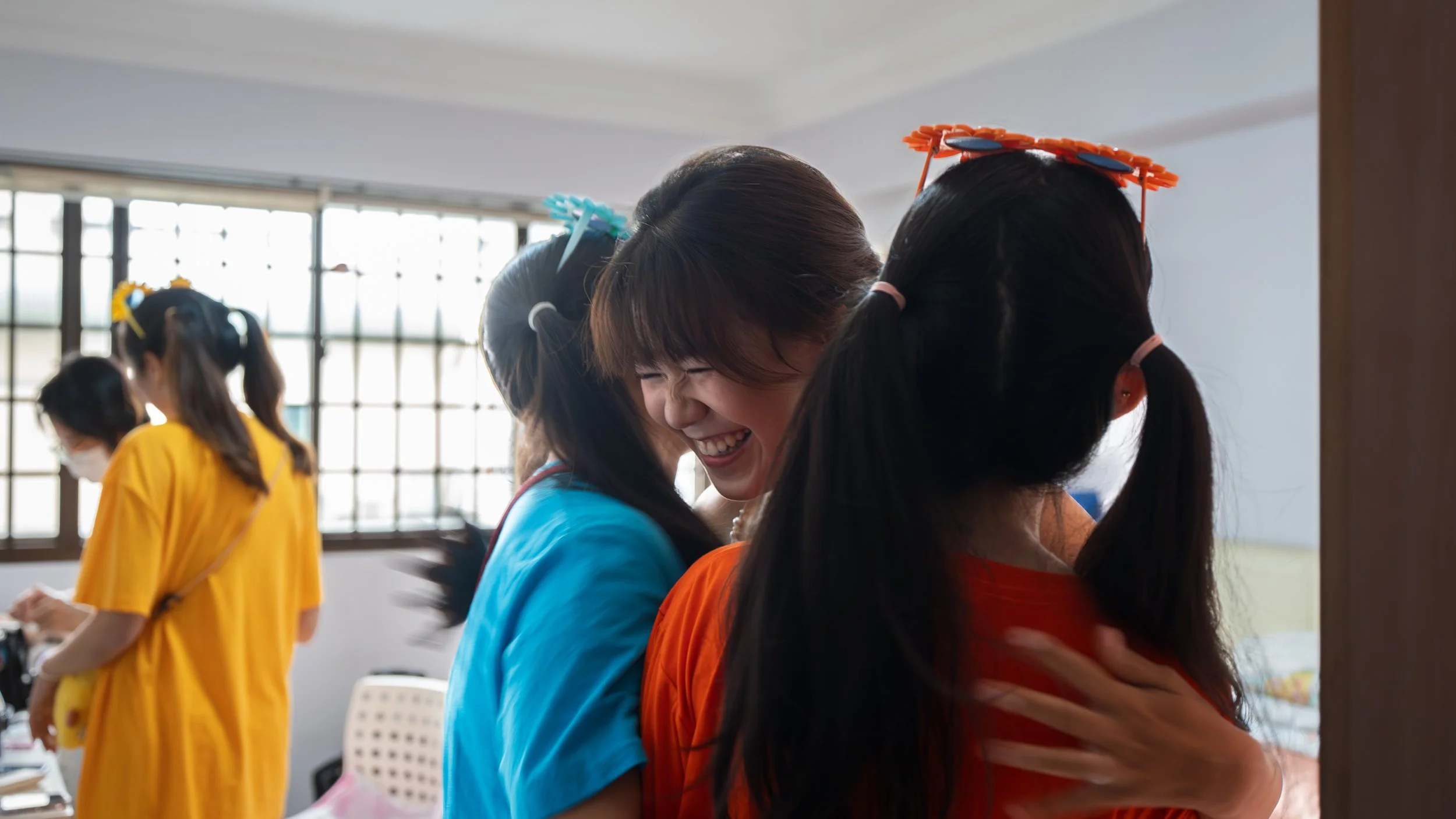 A group of young women wearing colorful T-shirts and hair accessories hugging and smiling in a bright room with large windows.