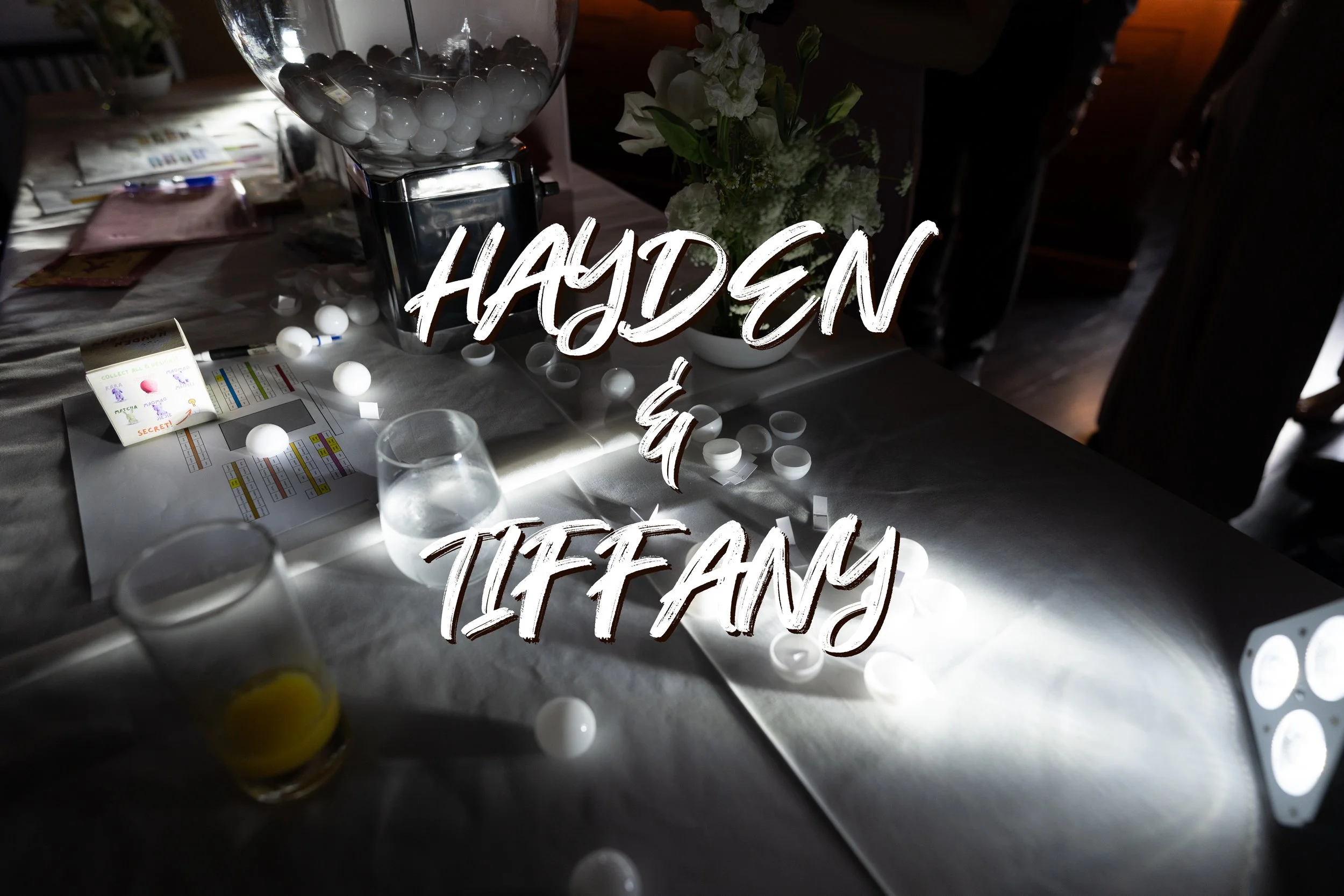 A table with a gumball machine, scattered white gumballs, a glass of water, a glass of orange juice, a small box labeled 'Secret,' papers, and a flower arrangement. Text overlay reads 'Hayden & Tiffany'.