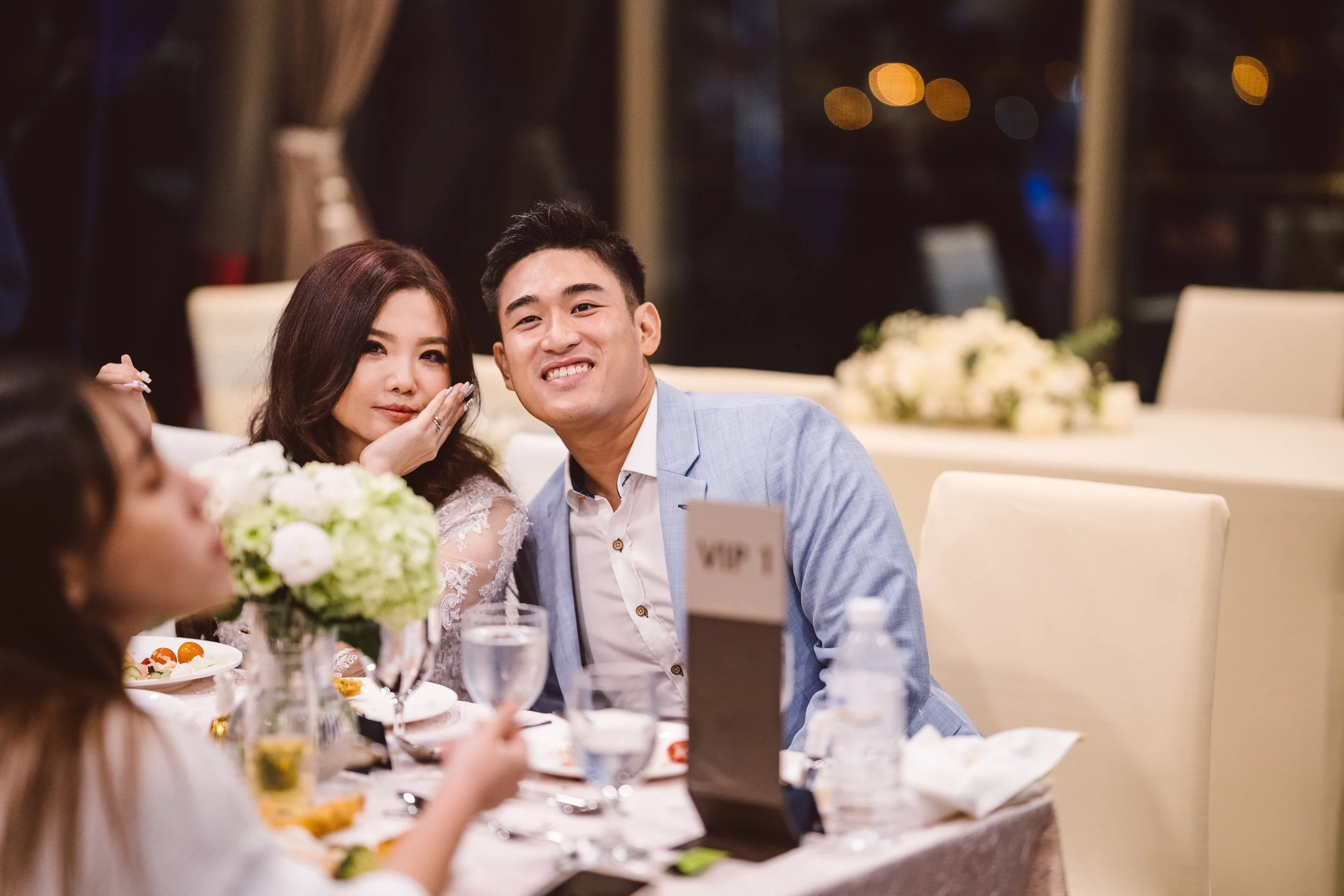 A group of people at a wedding reception, with a woman in a white lace dress making a cute face and resting her face on her hand, and a smiling man in a light blue suit sitting nearby, surrounded by flowers and table settings.