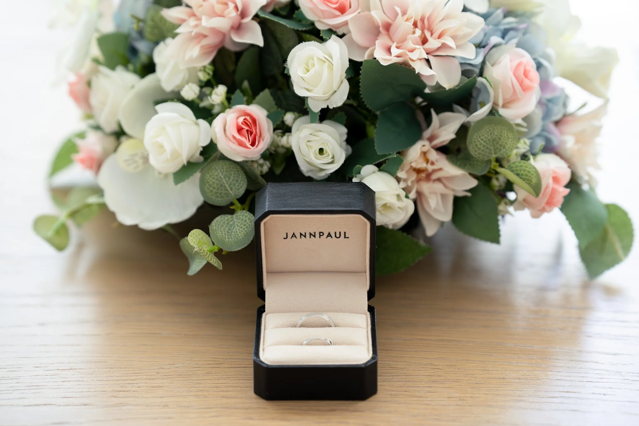Open black jewelry box with rings inside, placed on a wooden surface in front of a bouquet of pink and white flowers.