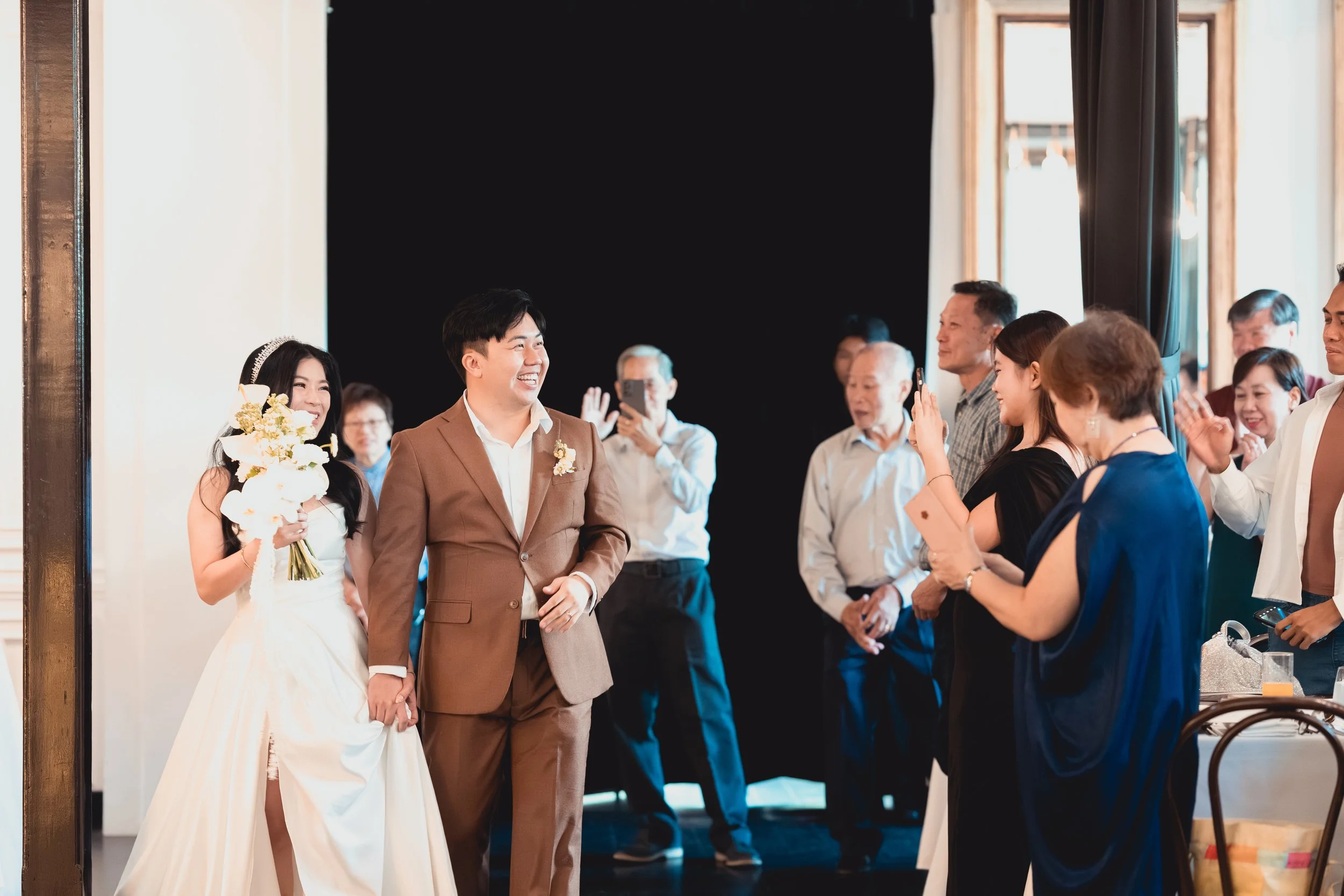 A joyous wedding reception with a bride and groom holding hands as they walk into a room filled with smiling guests. The bride is wearing a white wedding dress and holding a bouquet of flowers, while the groom is in a brown suit. Guests are clapping,