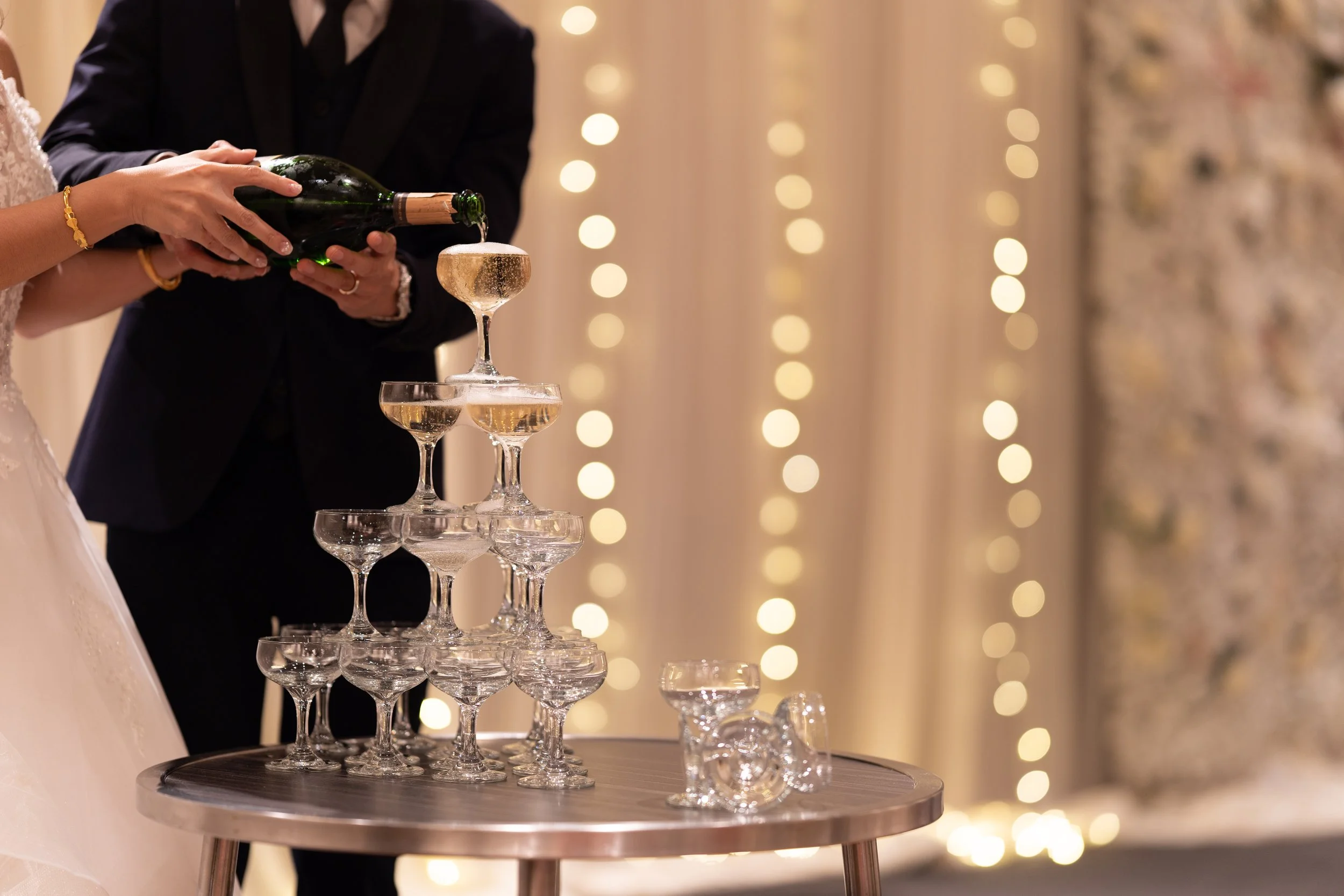Couple pouring champagne into a pyramid of glasses at a celebration event, with bokeh lights in the background.