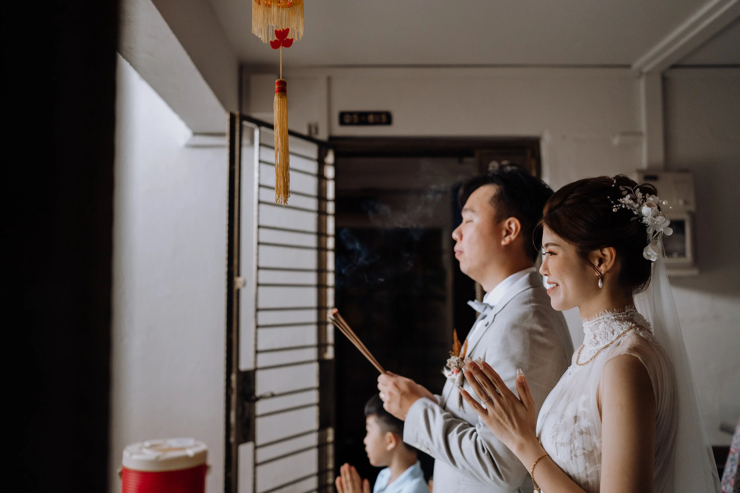 A bride and groom participating in a wedding ceremony, praying with their eyes closed, in an indoor setting, with a young boy in the background also praying.