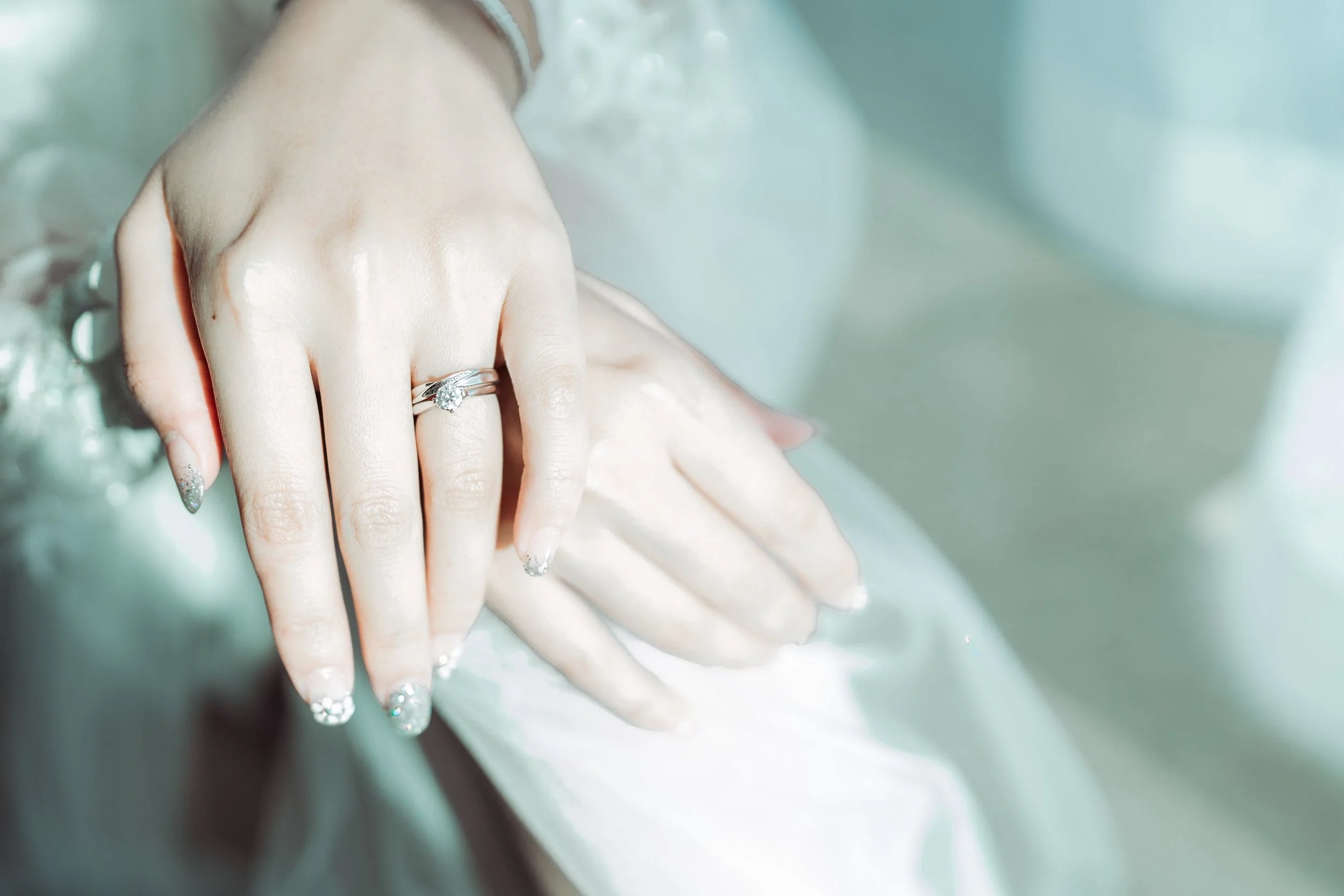 Close-up of a woman's hand with a diamond engagement ring, showing a manicure with sparkly tips, resting on a satin fabric with water drops.