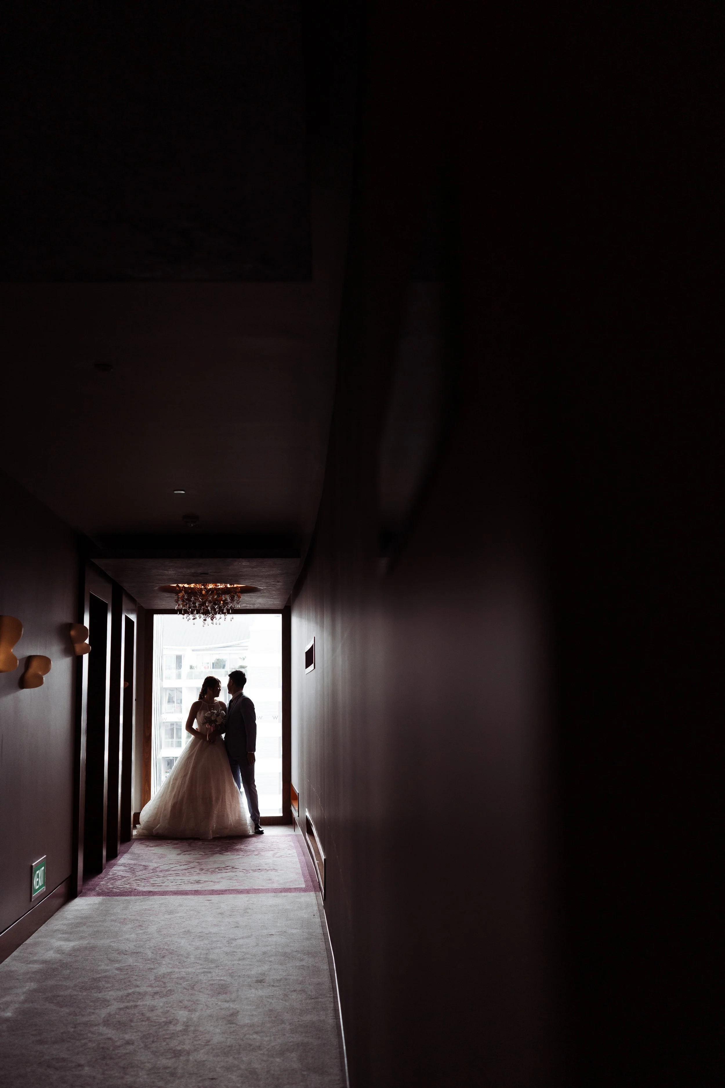 Silhouettes of a bride and groom holding hands in a corridor with a chandelier at the ceiling and a large window at the end of the hall.
