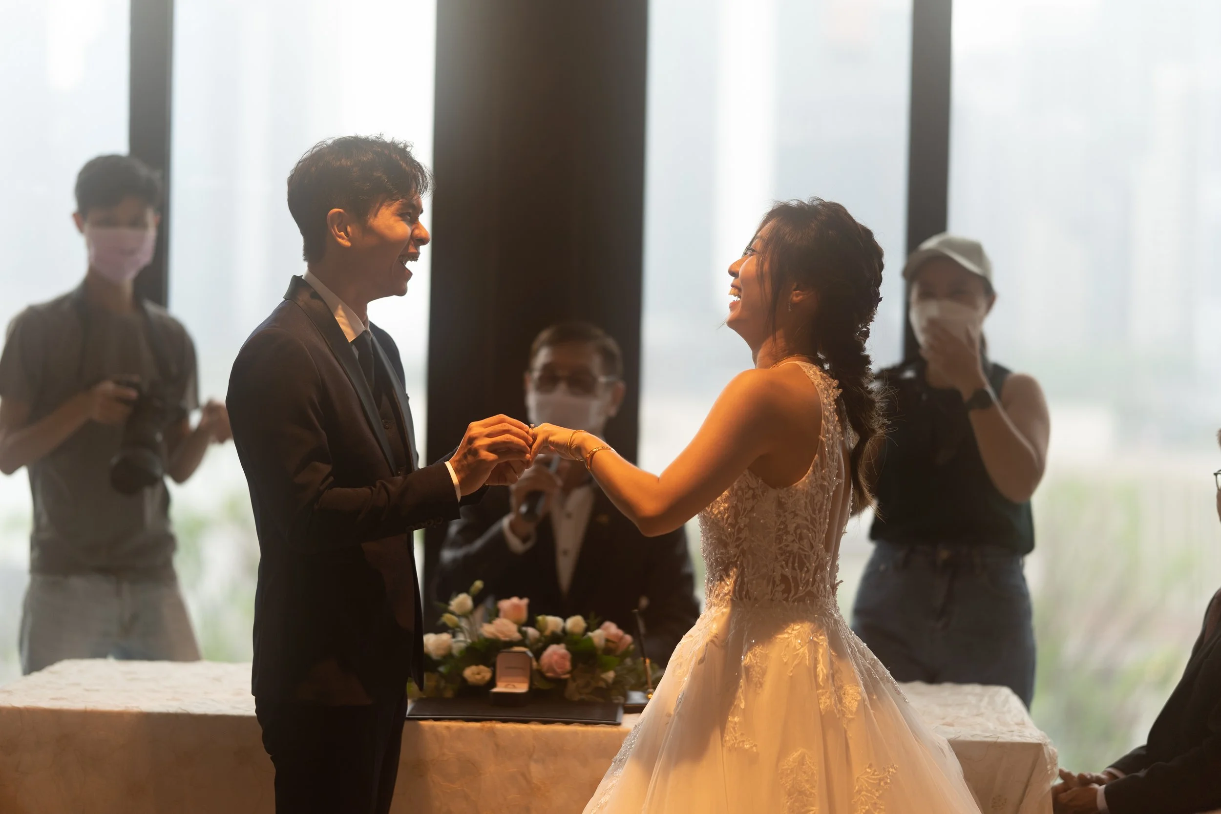A bride and groom smiling and holding hands during their wedding ceremony indoors, with wedding photographers and guests in the background.