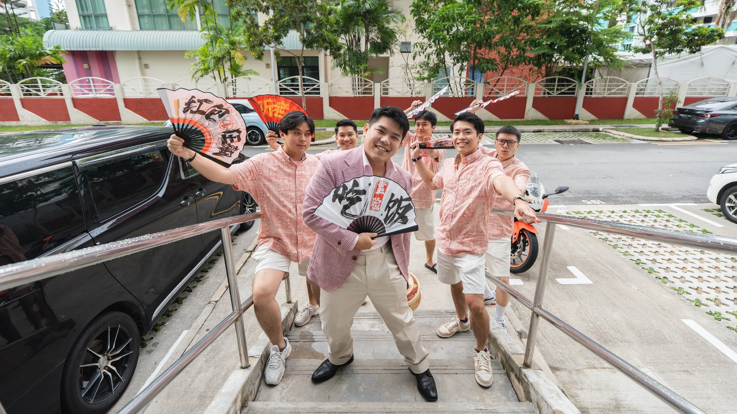 A group of six young men outdoors, smiling, holding colorful fans, and posing on a small set of steps. They are dressed in casual light-colored clothes, with some wearing matching pink patterned shirts and white shorts. There is a black car parked be