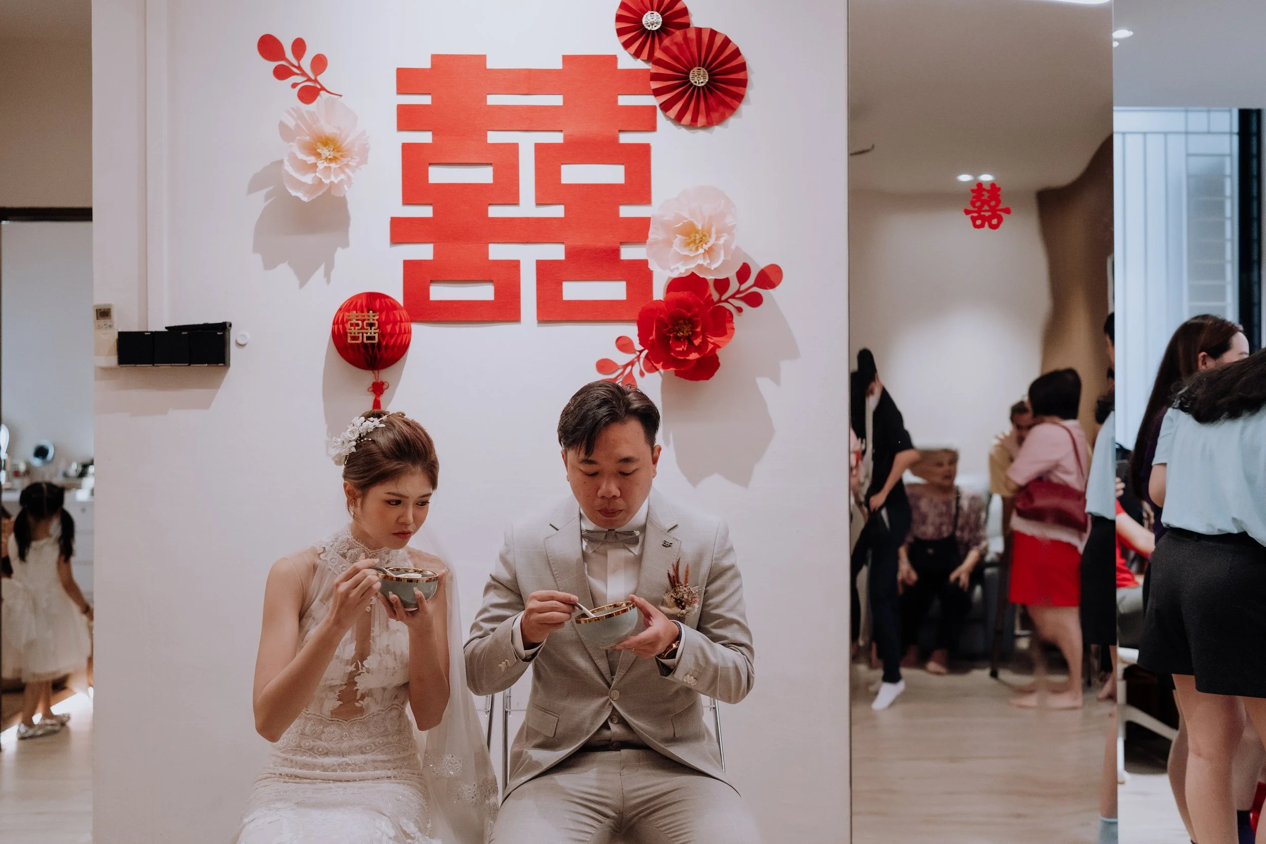 A bride and groom dressed in wedding attire sitting with bowls, participating in a traditional tea ceremony at a wedding, with festive decorations on the wall behind them including a large red double happiness symbol, paper lanterns, and paper flower