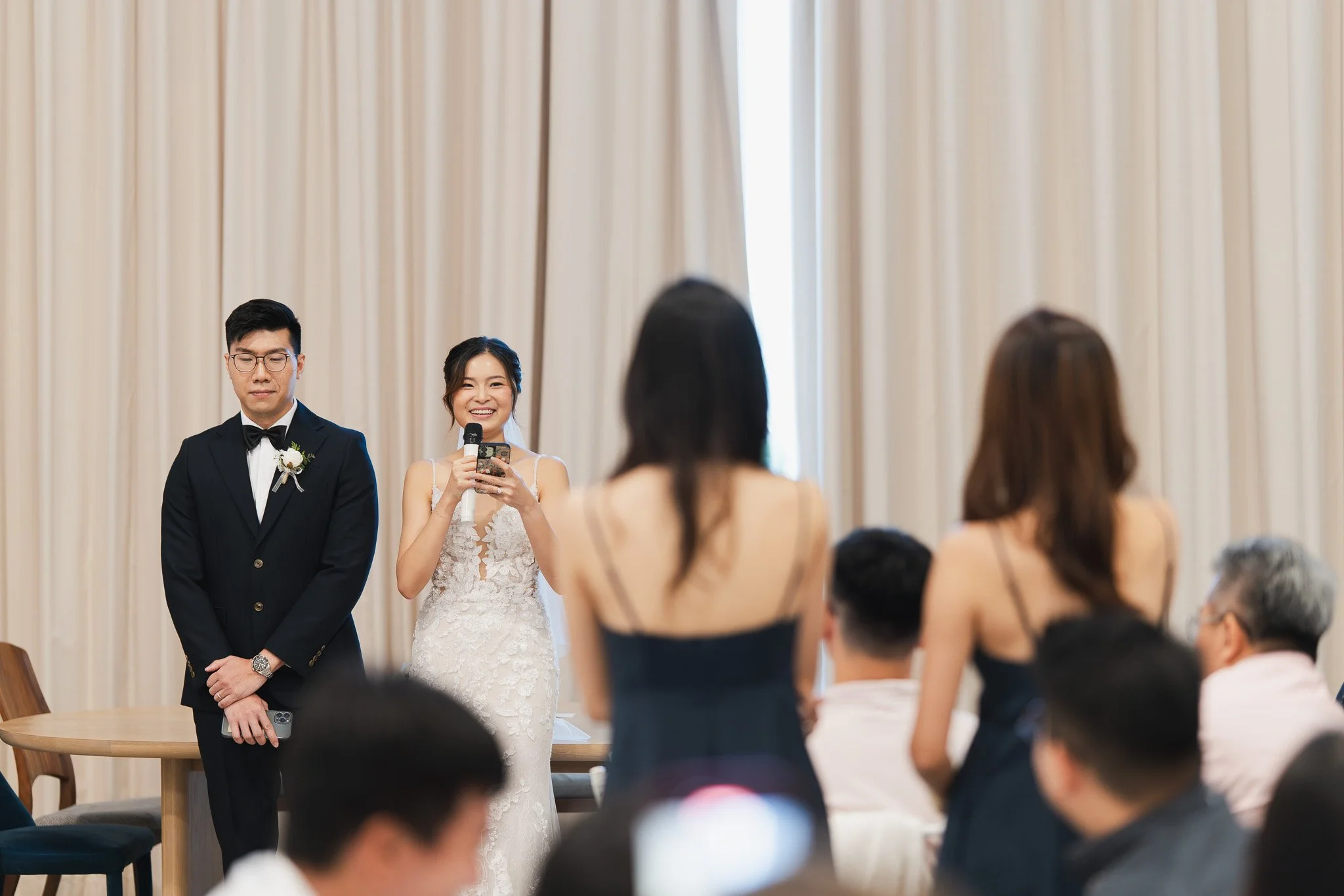 A wedding reception with a bride and groom standing in front of seated guests. The bride is smiling and holding a microphone and a phone, while the groom stands beside her with eyes closed, wearing a tuxedo. The background features beige curtains.