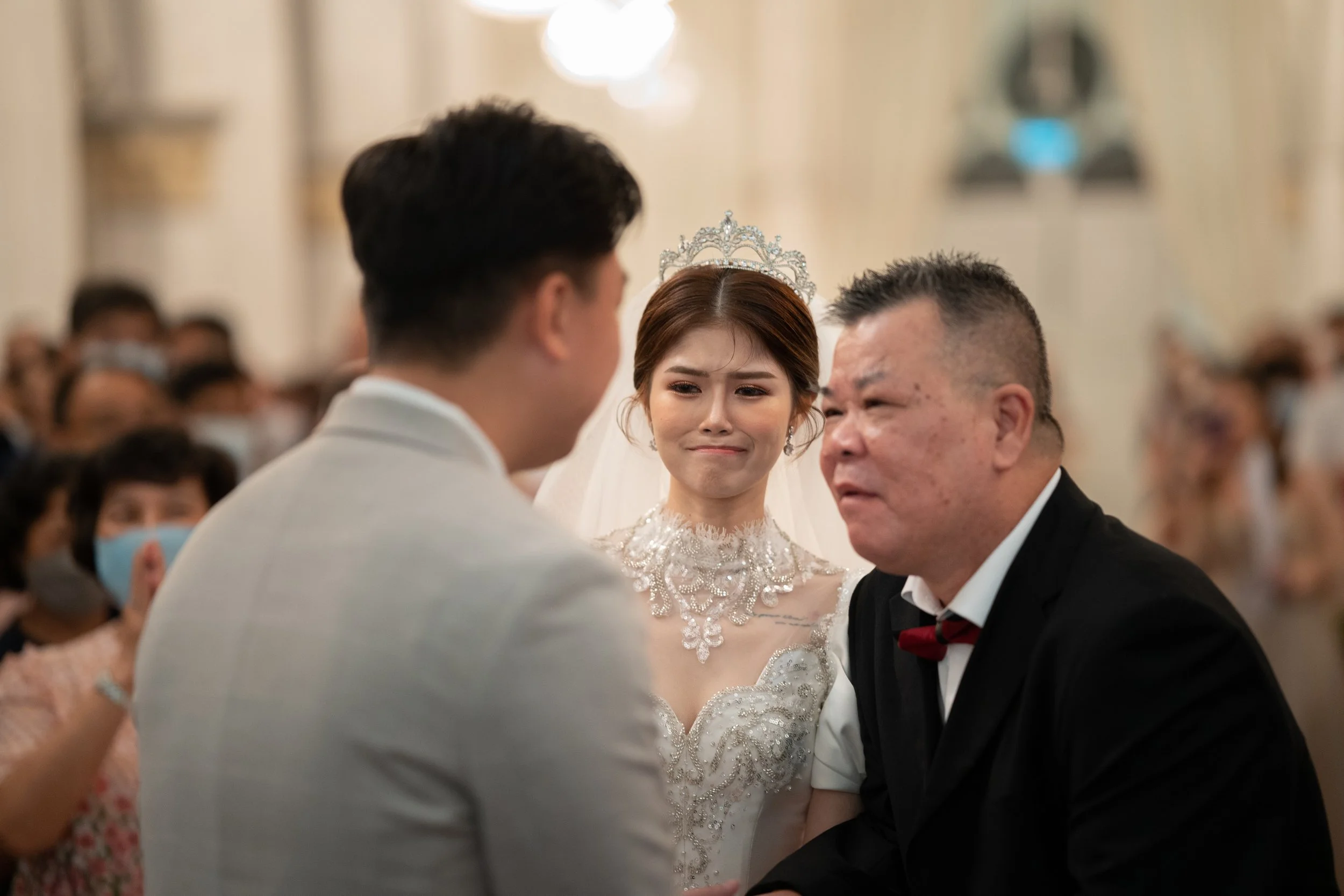 A bride with a tiara and wedding dress appears to be crying while looking at a groom during a wedding ceremony. An older man, possibly the father of the bride, is also present.