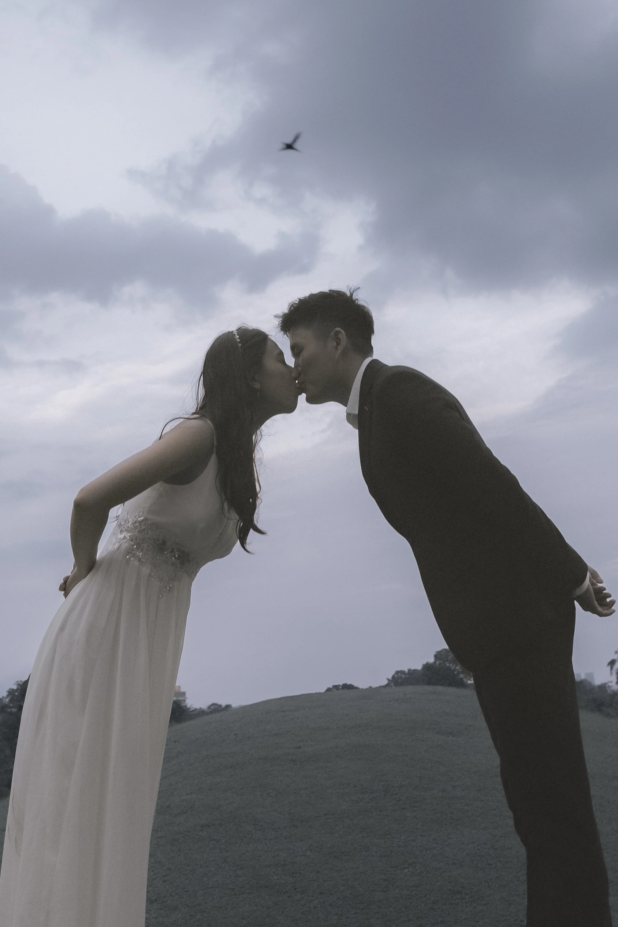 A couple dressed in wedding attire, a woman in a white dress and a man in a black suit, leaning in for a kiss outdoors during cloudy weather.