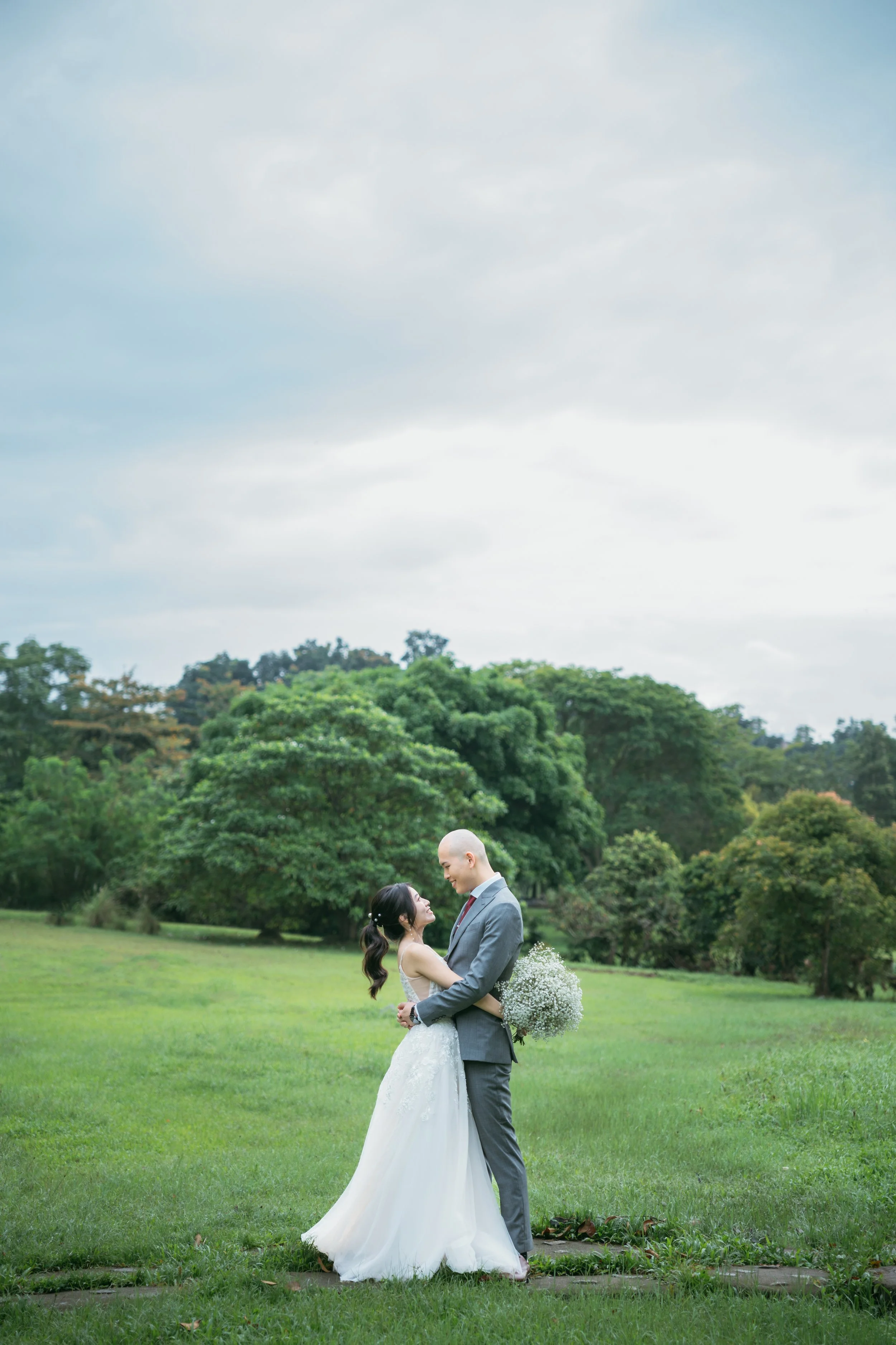 A bride and groom standing embrace in a lush green park, surrounded by trees under a cloudy sky during daytime.