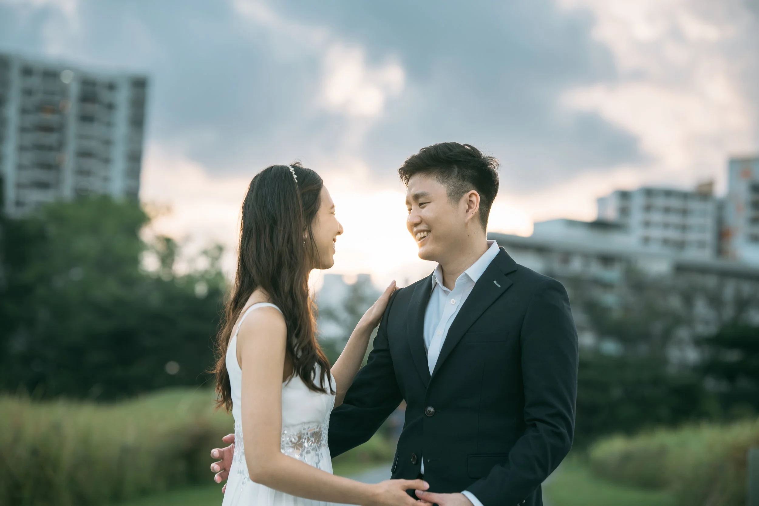 A couple dressed in formal attire, smiling and touching each other affectionately outdoors with city buildings in the background, during sunset.