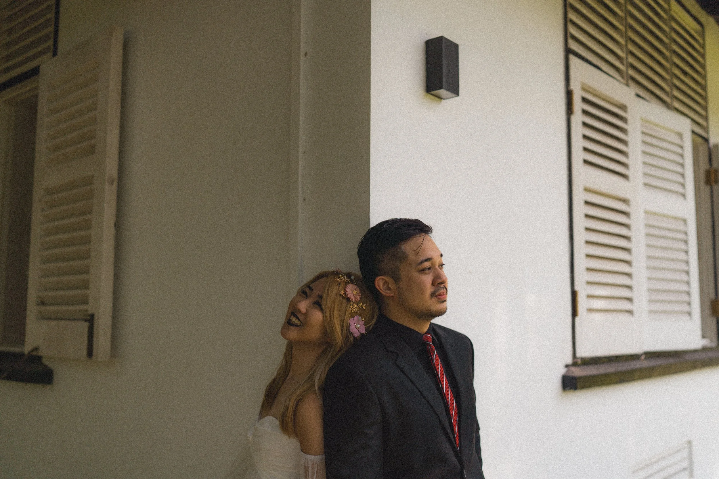 A man in a black suit and red tie stands with his back to a white wall, and a woman with long, blond hair and floral accessories, smiling with her head resting on his shoulder, standing against the same wall.