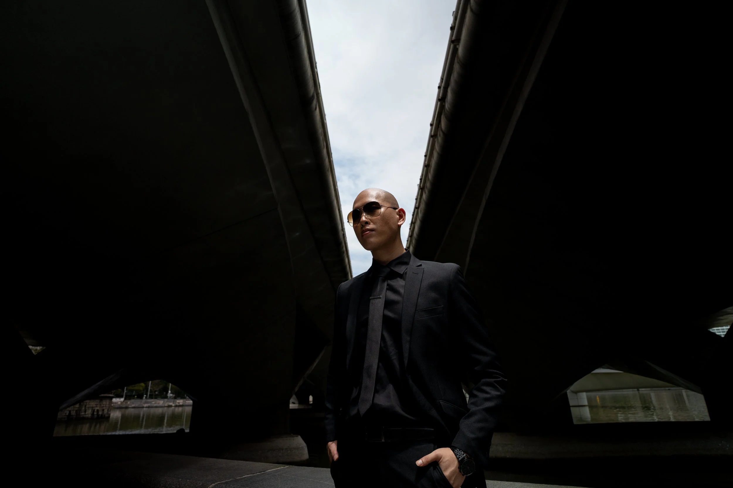 Man in black suit and sunglasses under a bridge with a cloudy sky