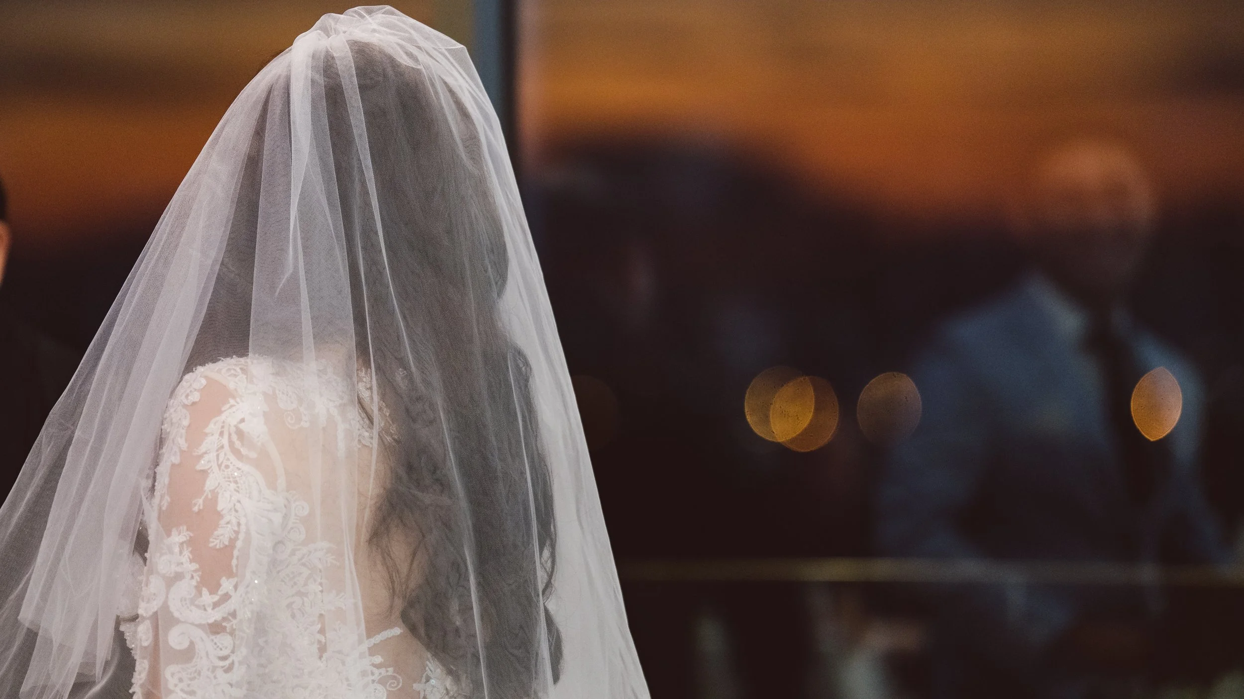 A woman in a wedding dress with lace details and a veil, standing in a dimly lit setting with blurred figures in the background.