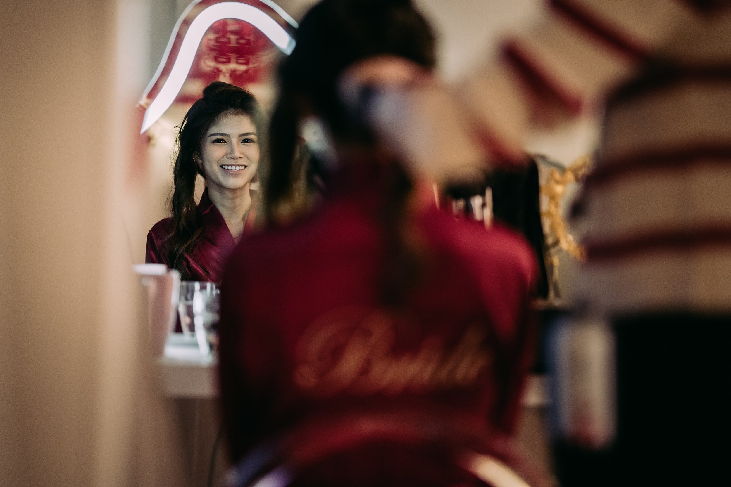 A young woman smiling at her reflection in a mirror, wearing a red satin robe, with blurred figures and long stacks of books visible in the foreground.