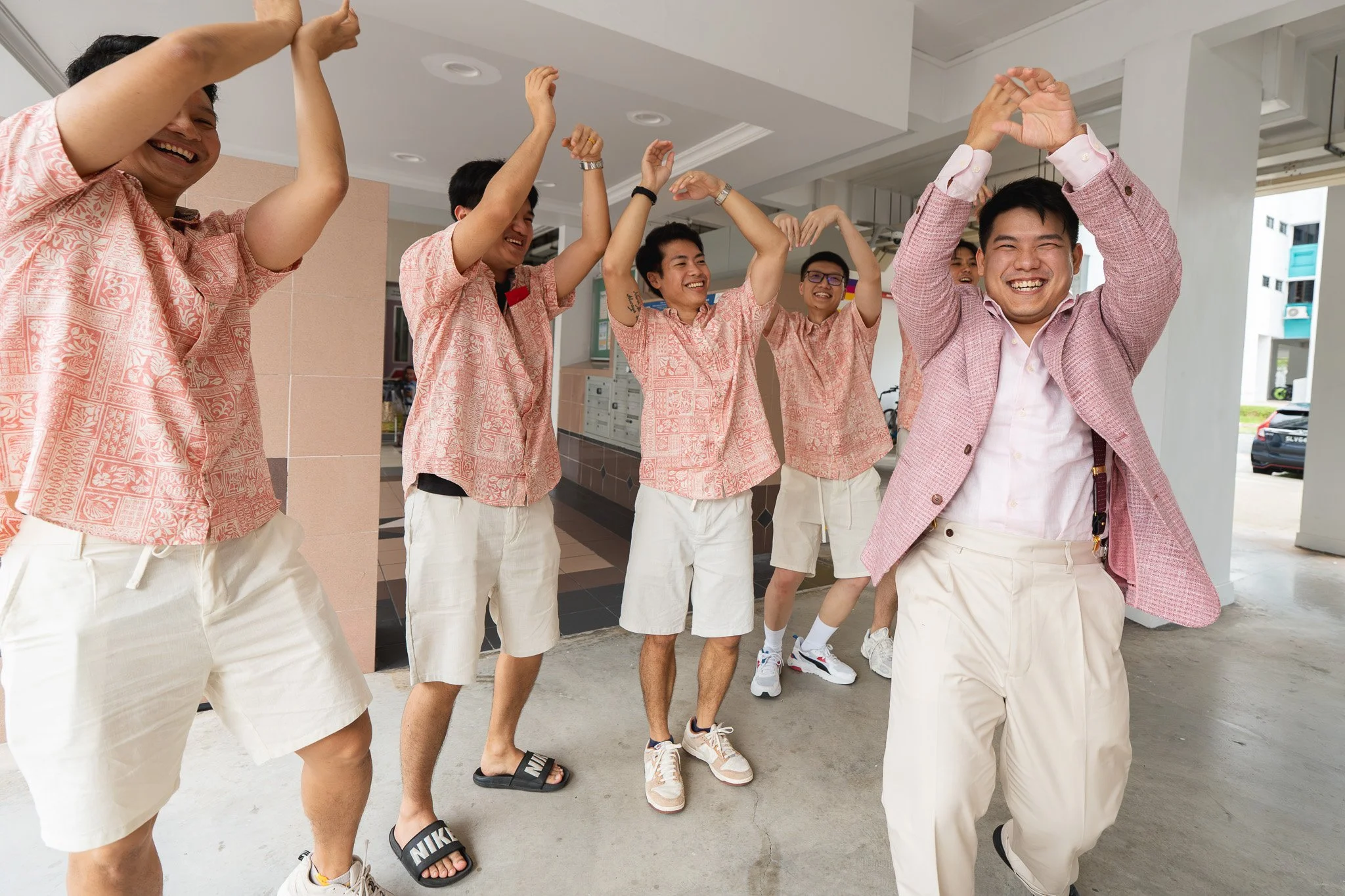 Group of young men wearing pink patterned shirts and light-colored shorts or trousers, smiling and dancing in a covered outdoor area.