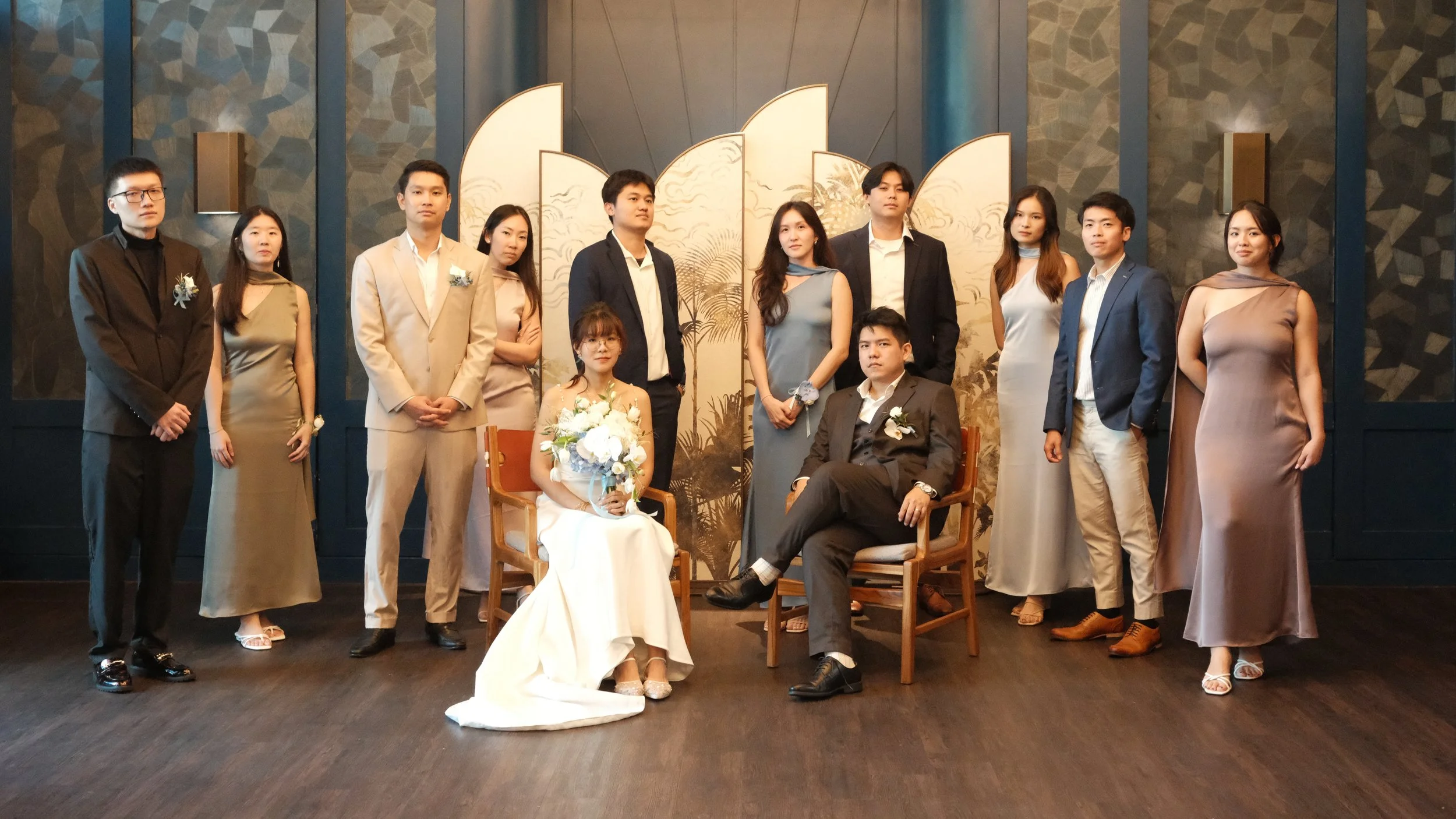 A group of people posing for a photo at a formal event, with a seated woman in a white dress holding a bouquet and a seated man in a dark suit, surrounded by others dressed in beige, black suits, and dresses, in front of decorative backdrop panels.