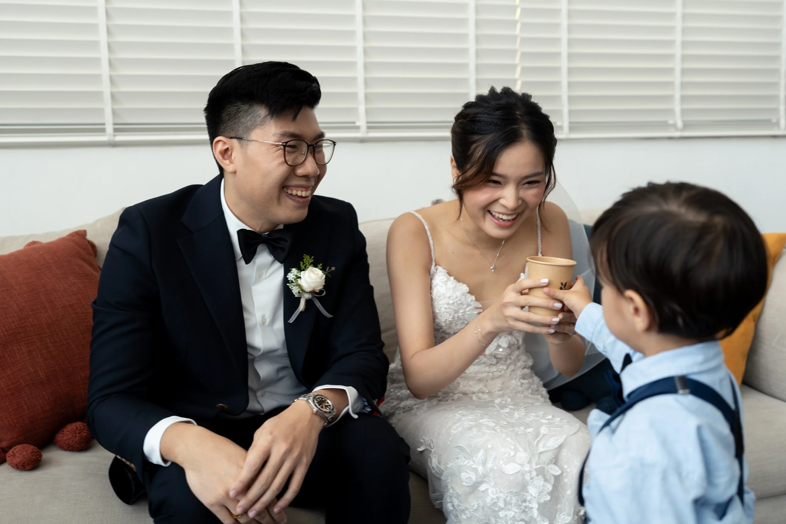 A wedding scene showing a bride and groom sitting on a sofa, smiling and sharing a moment with a small child who is offering a cup.