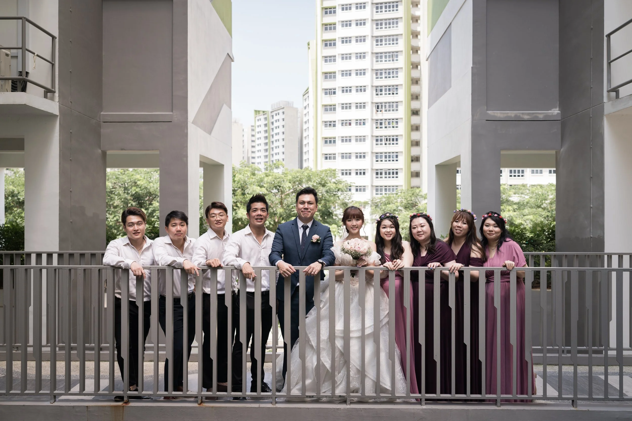 Group of people dressed for a wedding on a balcony in an urban area, with buildings and trees in the background.