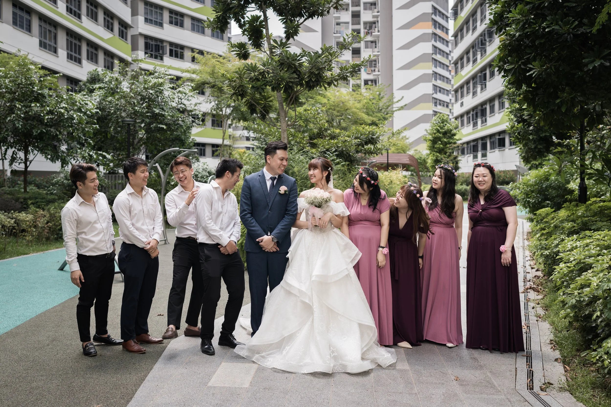 Bride and groom standing together with wedding party outdoors in an urban park, surrounded by tall apartment buildings and greenery.