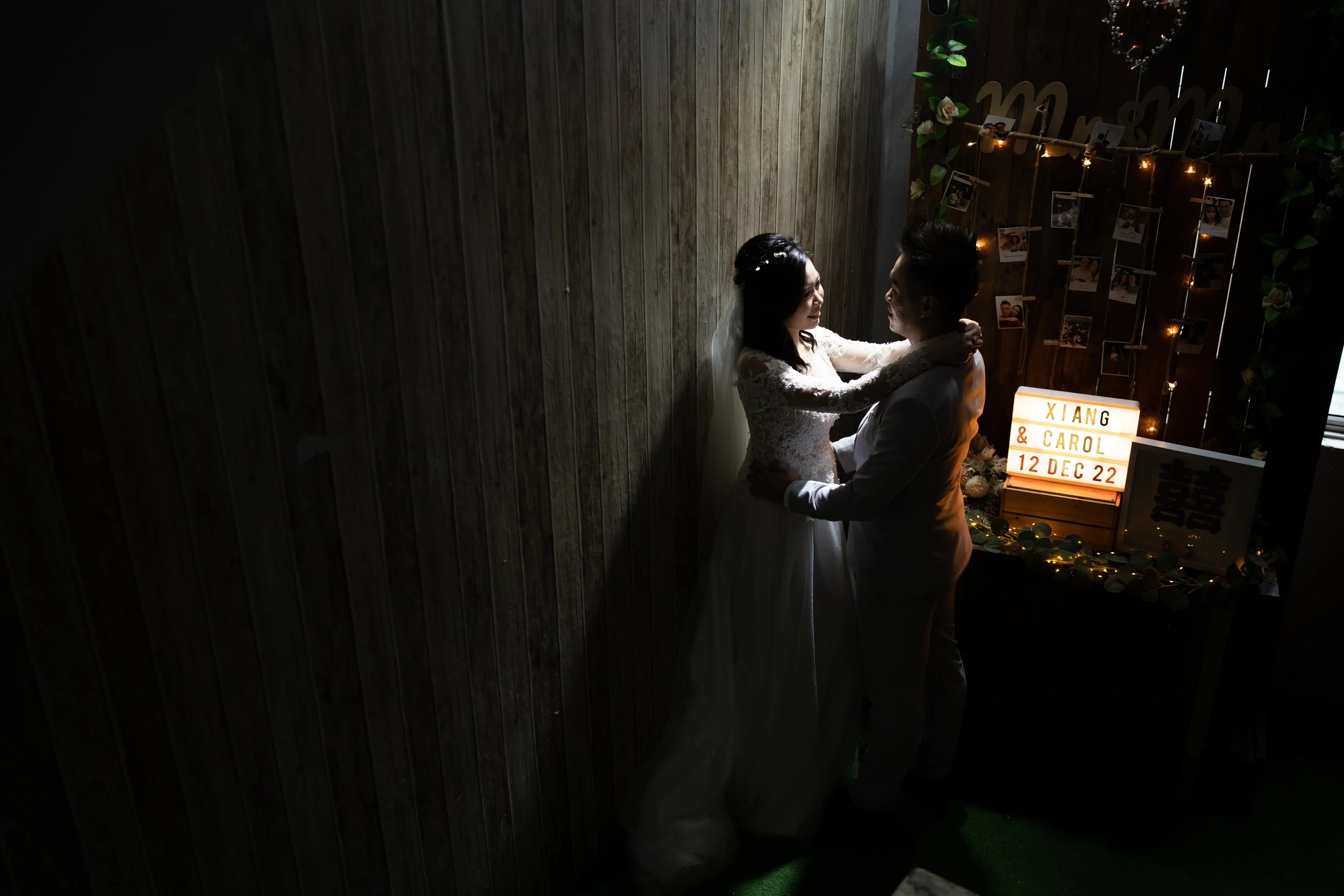 Bride and groom dancing in a dimly lit room, with a decorated background featuring a lightbox reading 'Xiang & Carol 12 Dec 22' and a collage of photos.