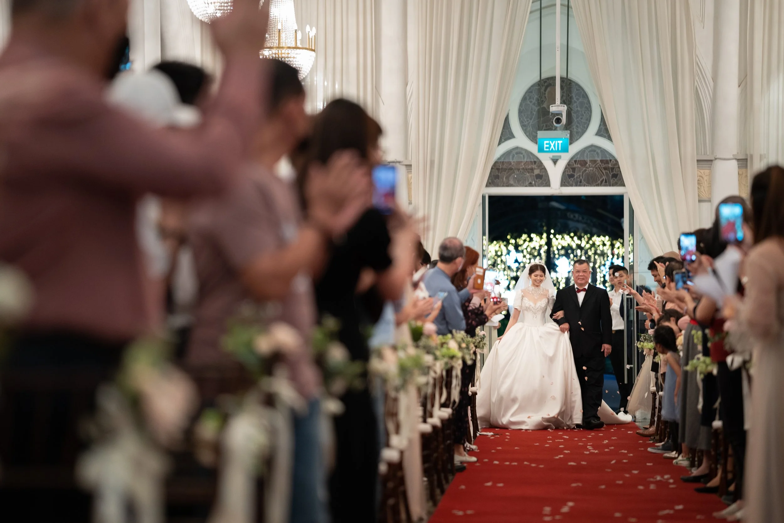A bride in a white wedding gown walking down the aisle with an older man in a tuxedo, surrounded by guests taking photos in a decorated wedding venue.