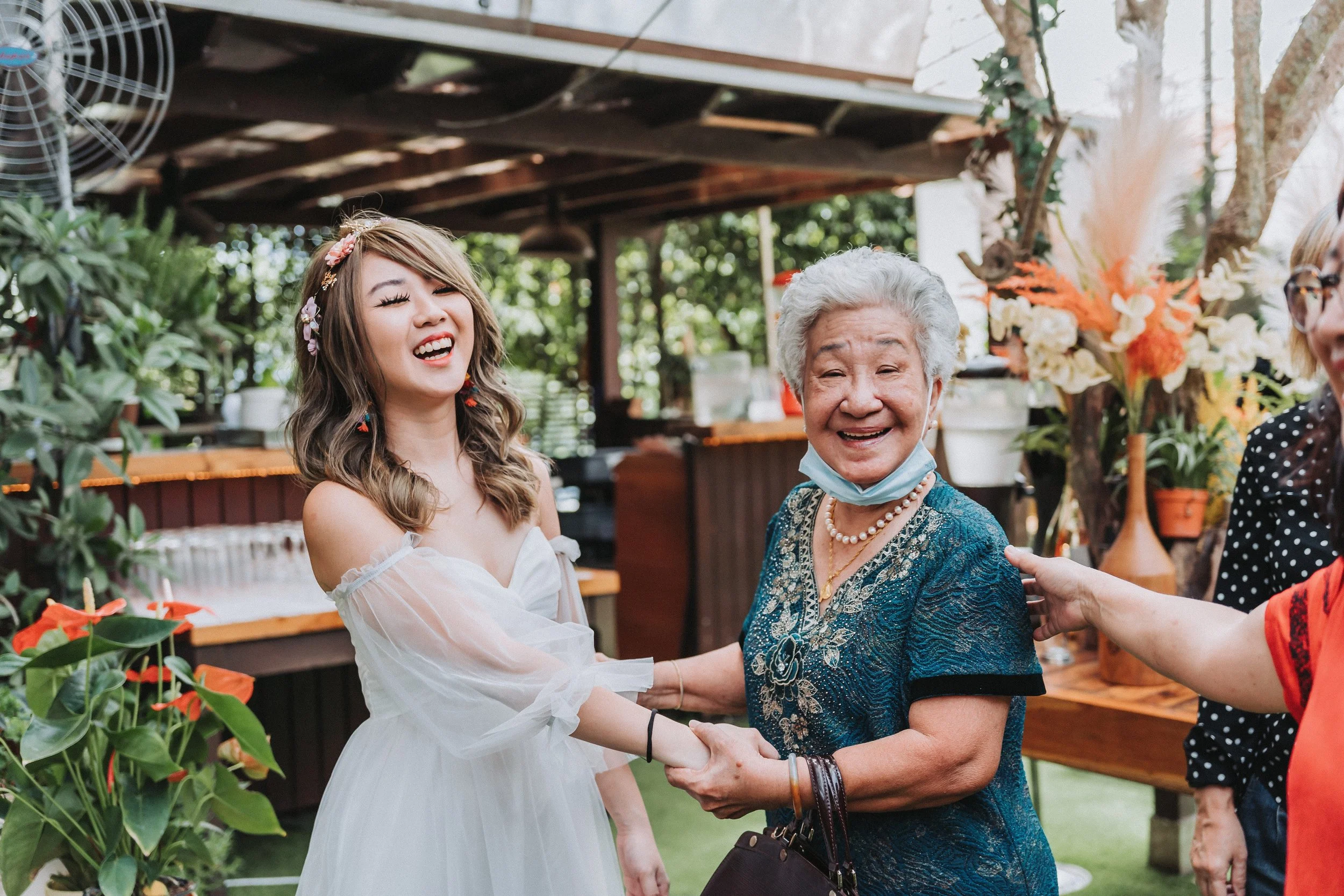 A young woman in a white dress and an elderly woman in a blue dress are smiling and holding hands at an outdoor gathering with flowers and greenery in the background.