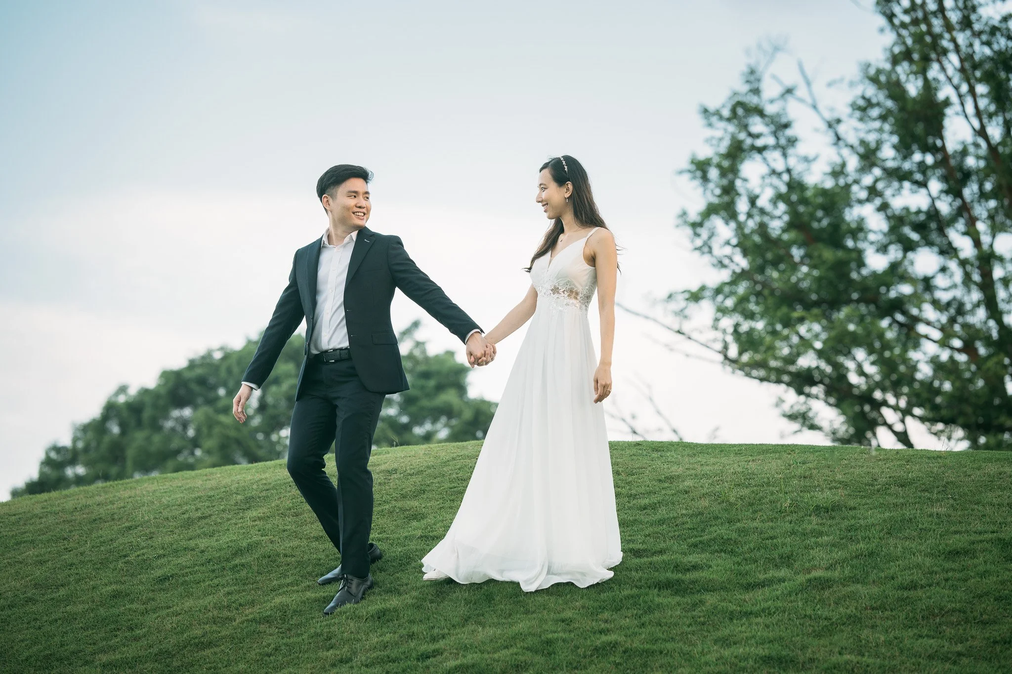 A smiling couple dressed in wedding attire holding hands on a grassy hill outdoors with trees in the background.