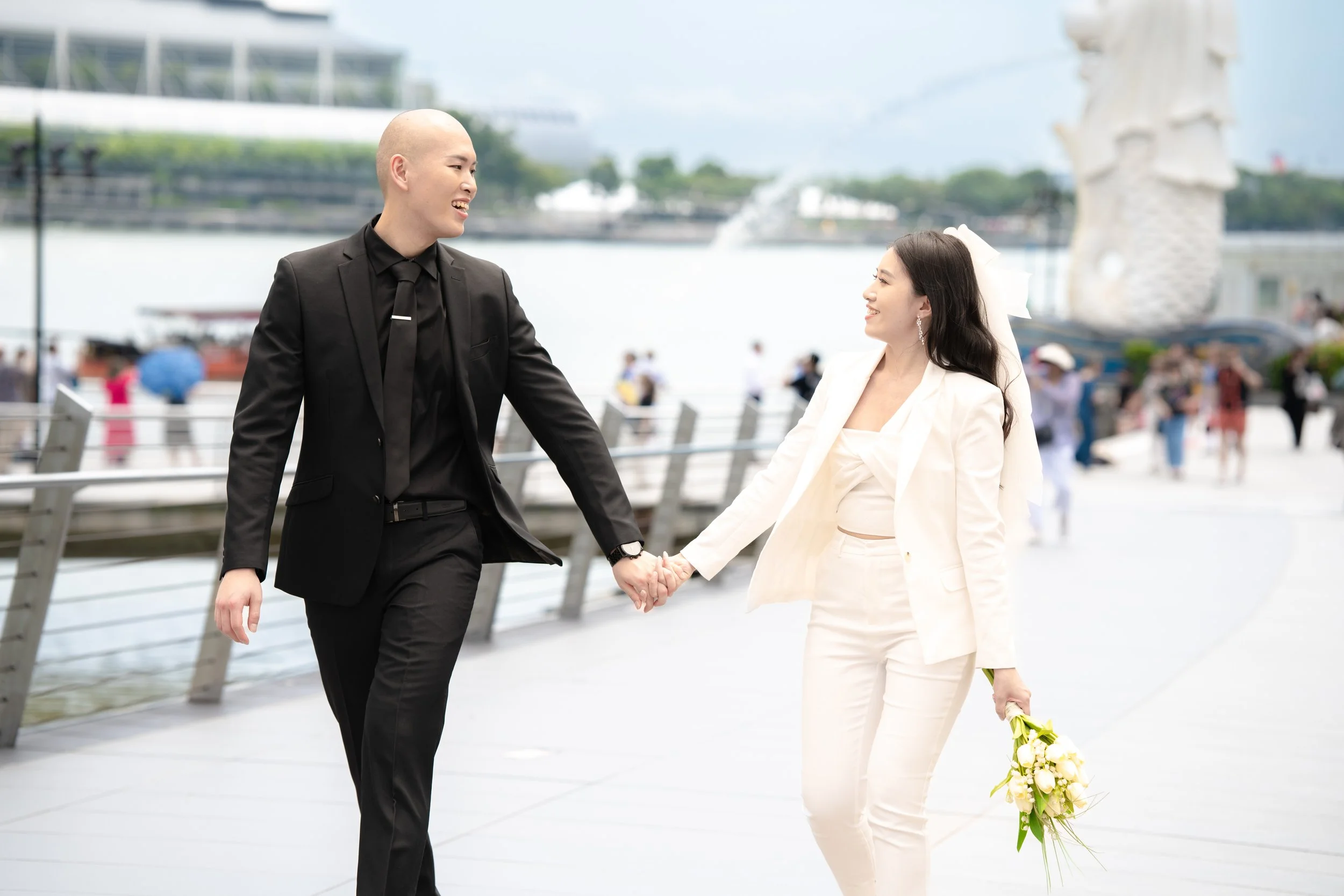 A couple dressed in formal wedding attire holding hands near a waterfront with the Merlion statue in Singapore in the background.