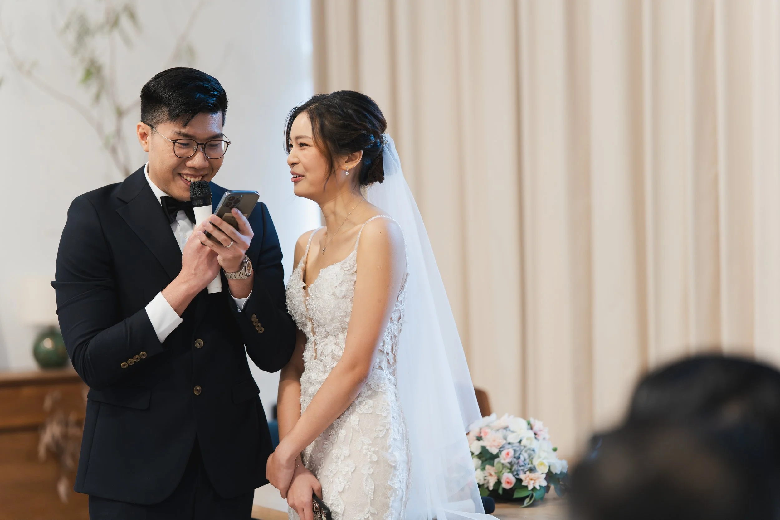 A bride and groom at their wedding ceremony, with the groom reading vows from a phone while holding a microphone, and the bride smiling.