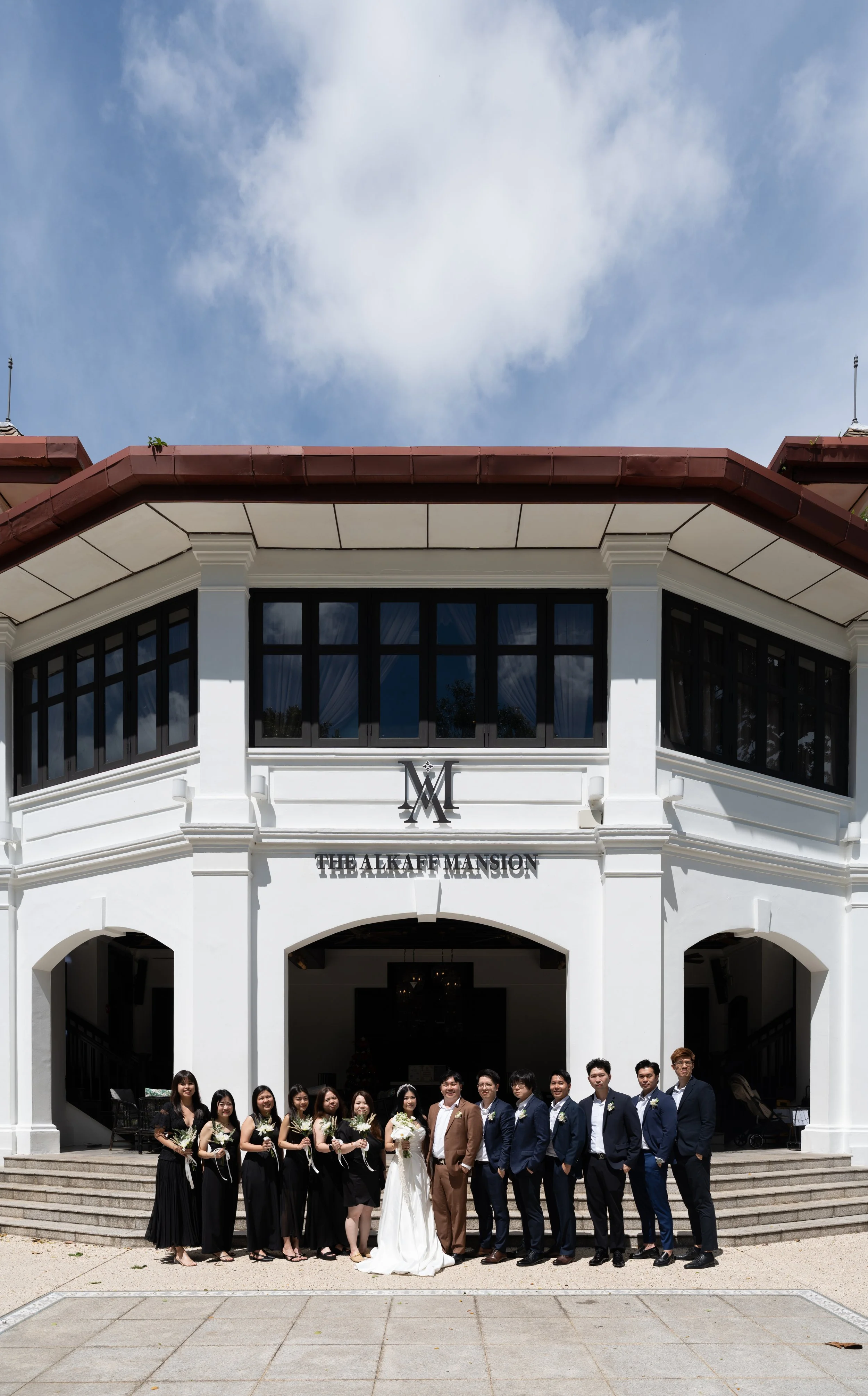 Group of people in wedding attire standing in front of a white mansion with black window frames and a sign that reads 'The Alkaef Mansion'