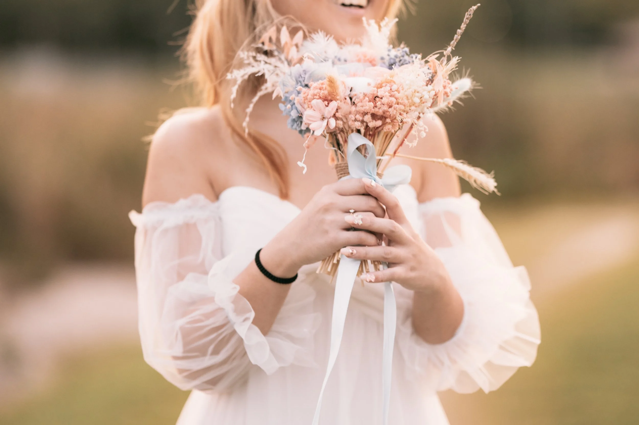 A woman in a white off-shoulder dress holding a bouquet of pastel-colored flowers, smiling outdoors.