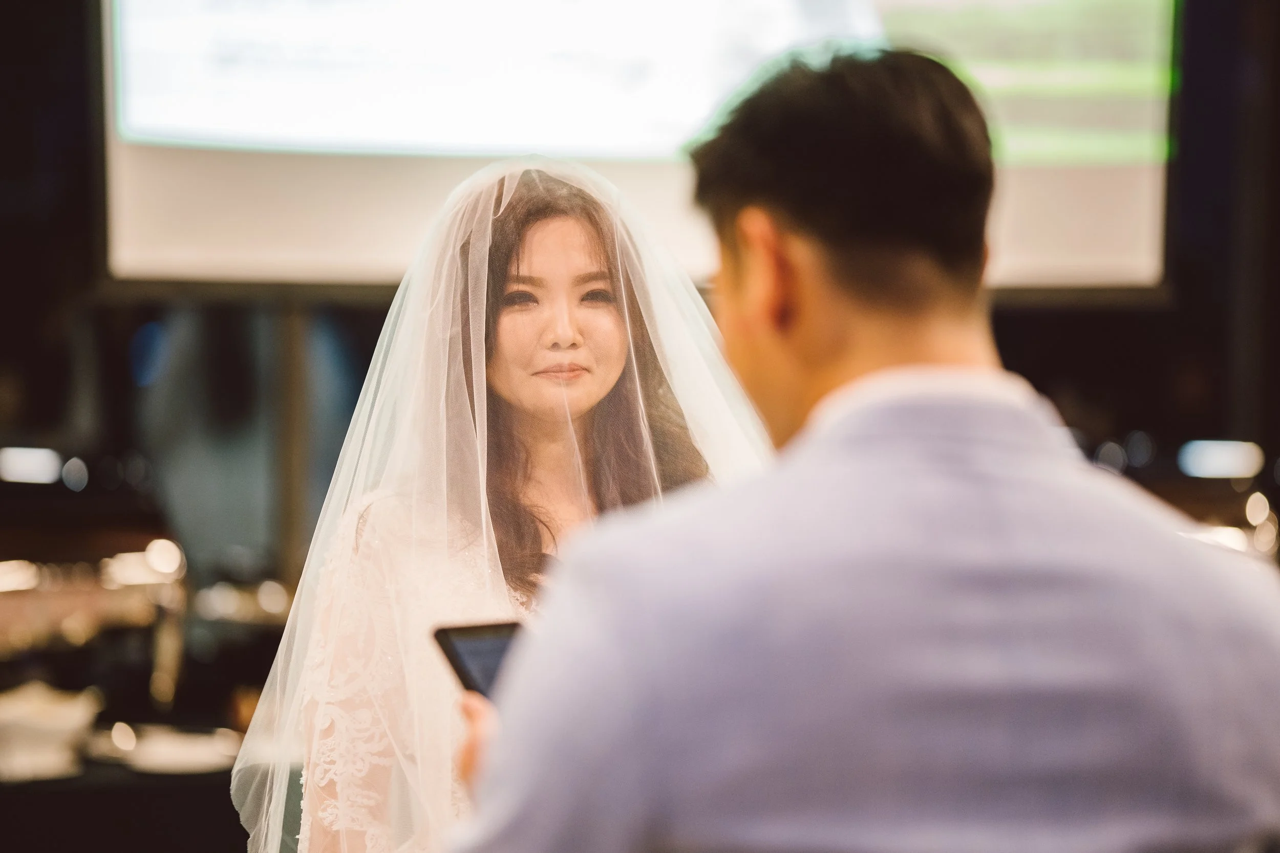 A woman in a wedding dress and veil smiling at a man in a suit who is using a smartphone, with a blurred certificate or paper in the background.